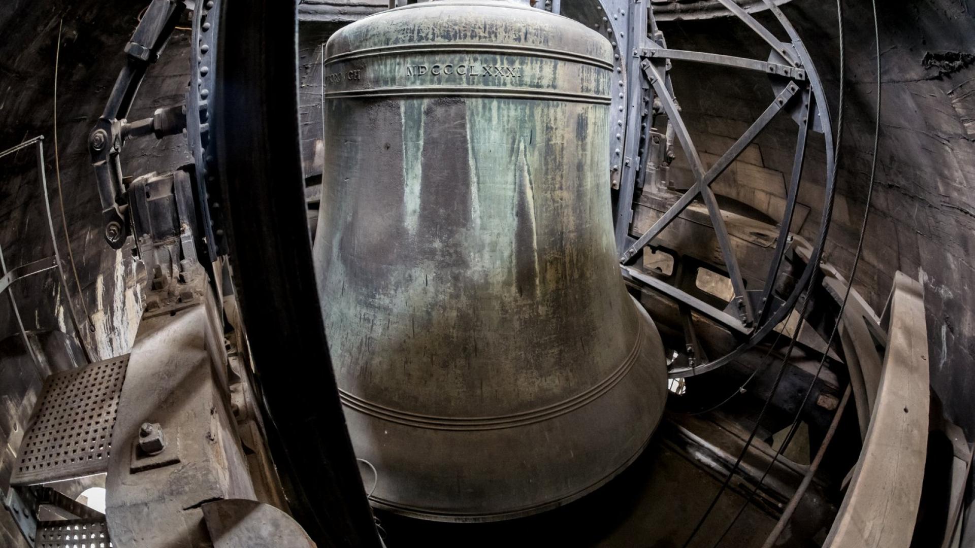 A huge cast bell, stained green in places, held in place by a large iron structure, inside a stone building