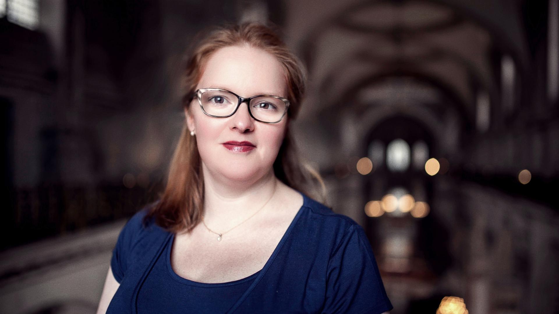 Carris, a white woman with auburn hair and glasses, stands in the cathedral, the lights of the nave and the decorative ceiling blurred in the background behind her.
