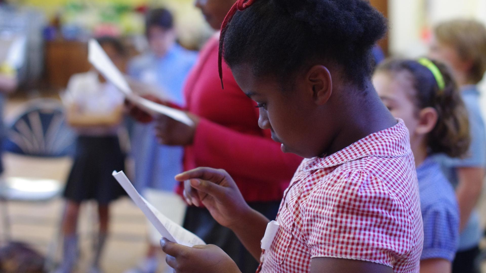 A young black girl in a red and white checked school summer dress is intently reading sheet music