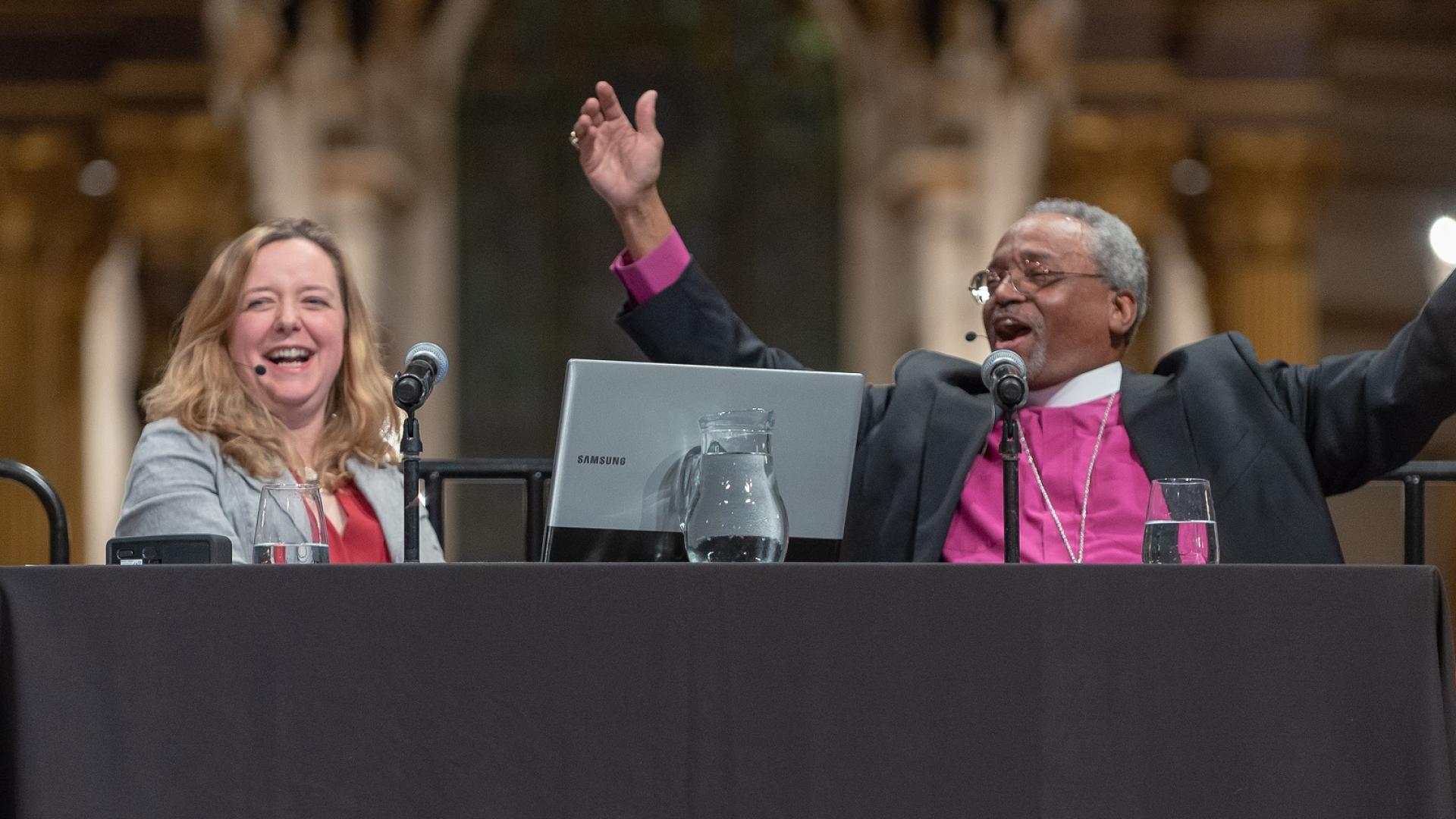 Bishop Curry, a black man with grey hair in a clerical collar purple shirt, on stage under the dome of the cathedral, smiling and arms outstretched, alongside a smiling Paula Gooder, a white woman with long blond hair in a grey jacket and red blouse.