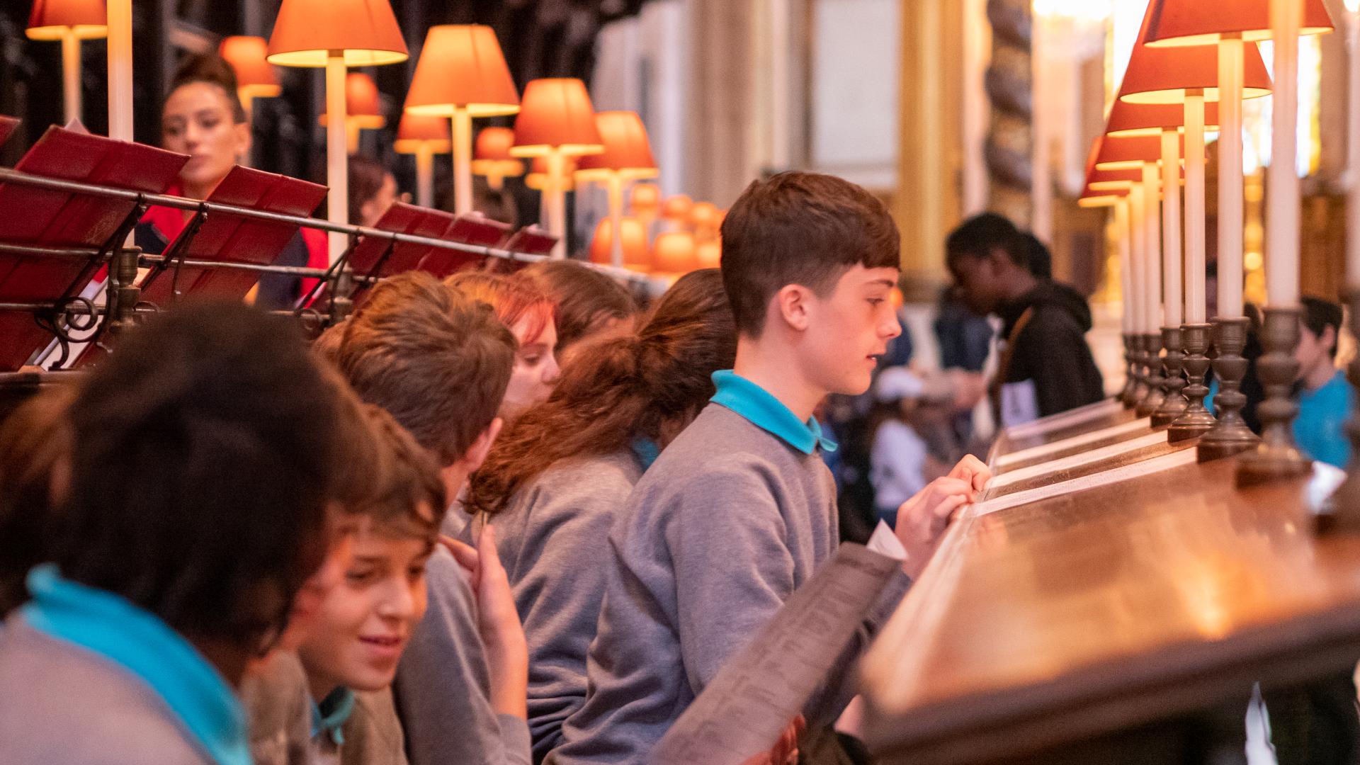 Secondary school age children in grey jumpers and blue polo shirts sit in the quire