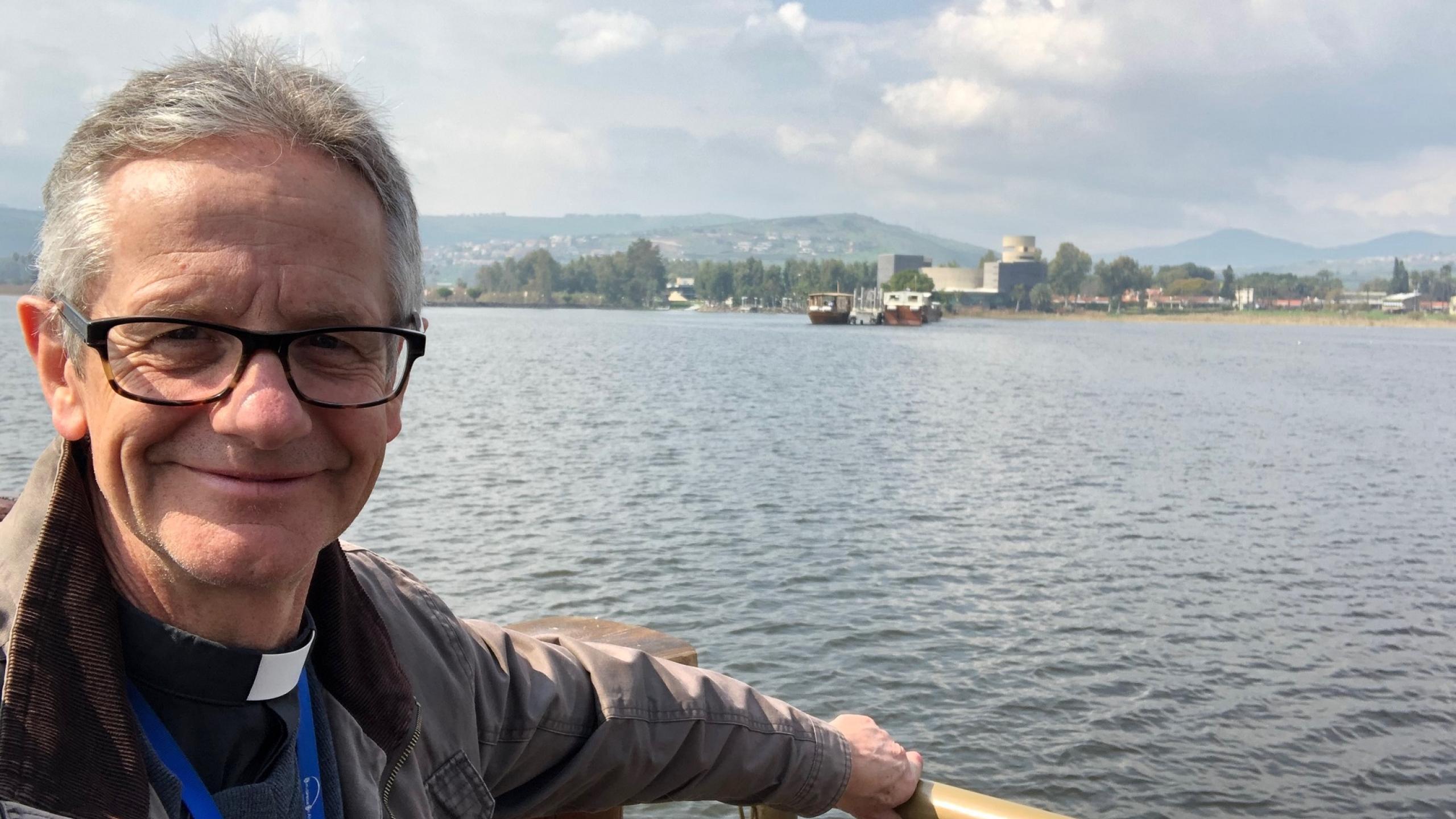 Richard is a white man with short grey hair and black rimmed glasses. He wears a clerical collar and black shirt under a casual light jacket and stands holding onto the wooden railing of a boat or a jetty on a lake. The shoreline in the distance has trees, and some buildings, and there are mountains in the distant background. The sky is pale blue with some clouds and the sun is shining on Richard.