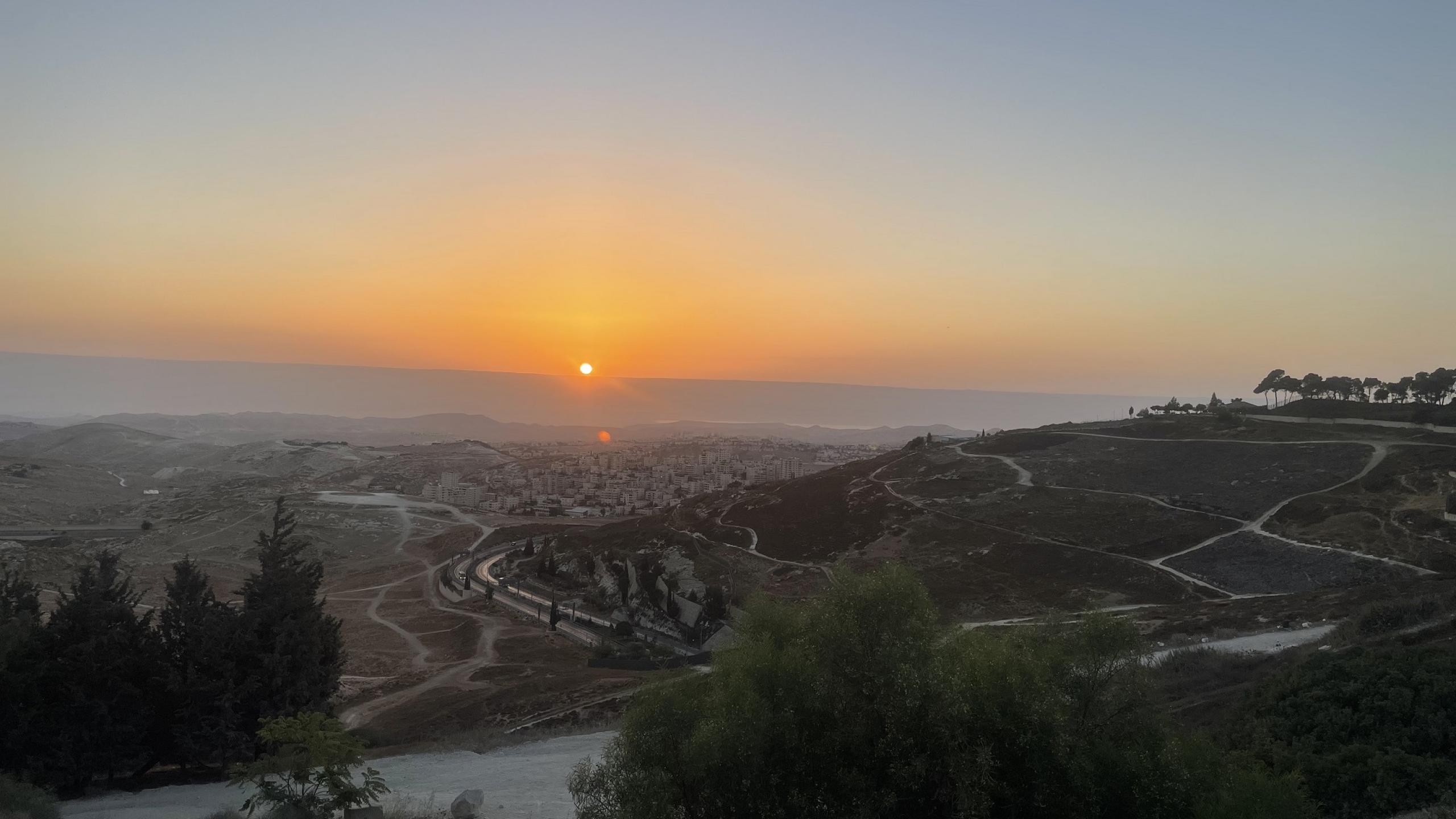 A view over the Jordan Valley from the Mount of Olives, looking down towards a distant town with the sun rising in an orange-tinged sky in the distance.