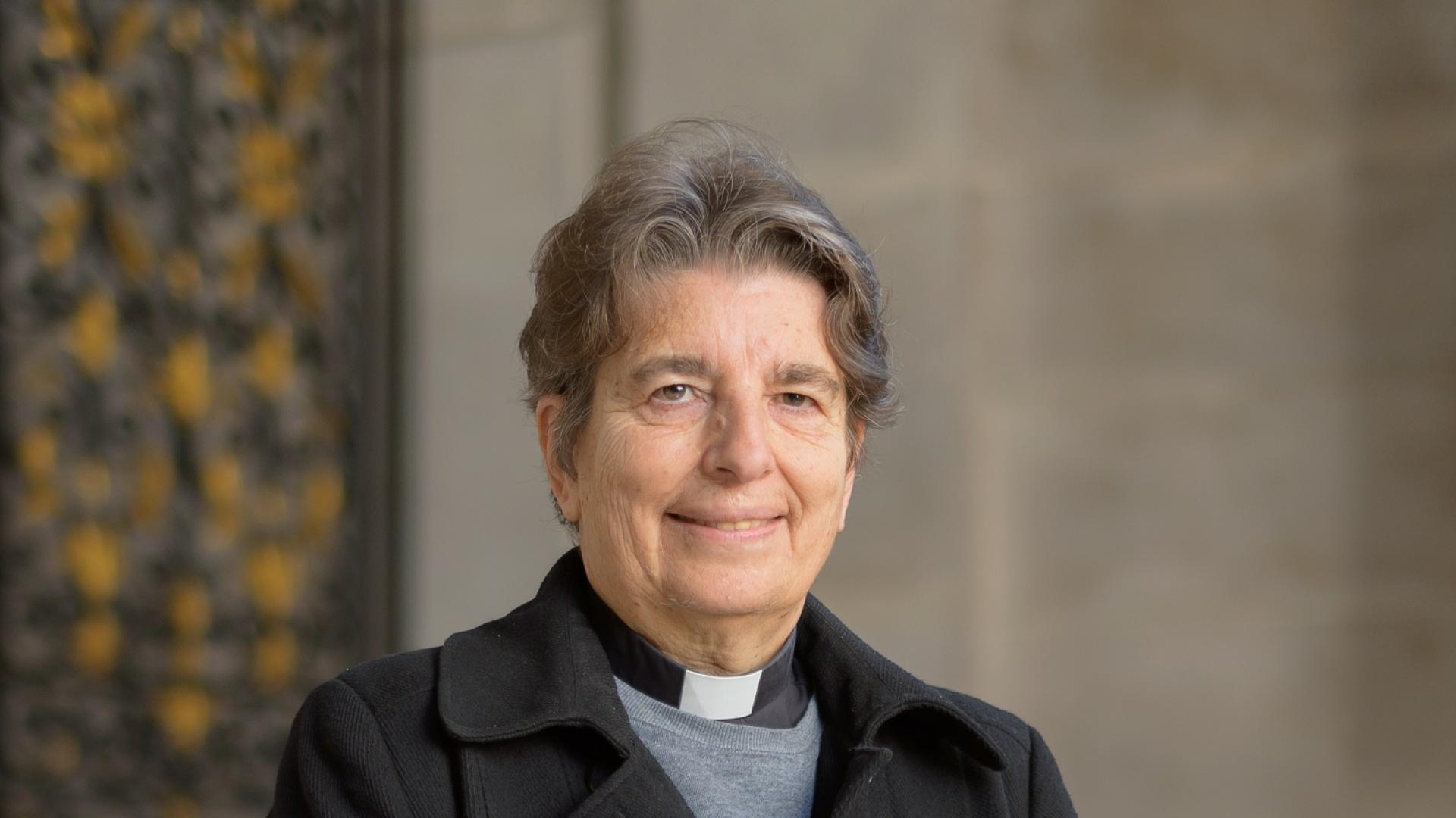 Paula is a white woman with short dark grey hair and wears a clerical collar under a grey shirt and black double breasted coat. 