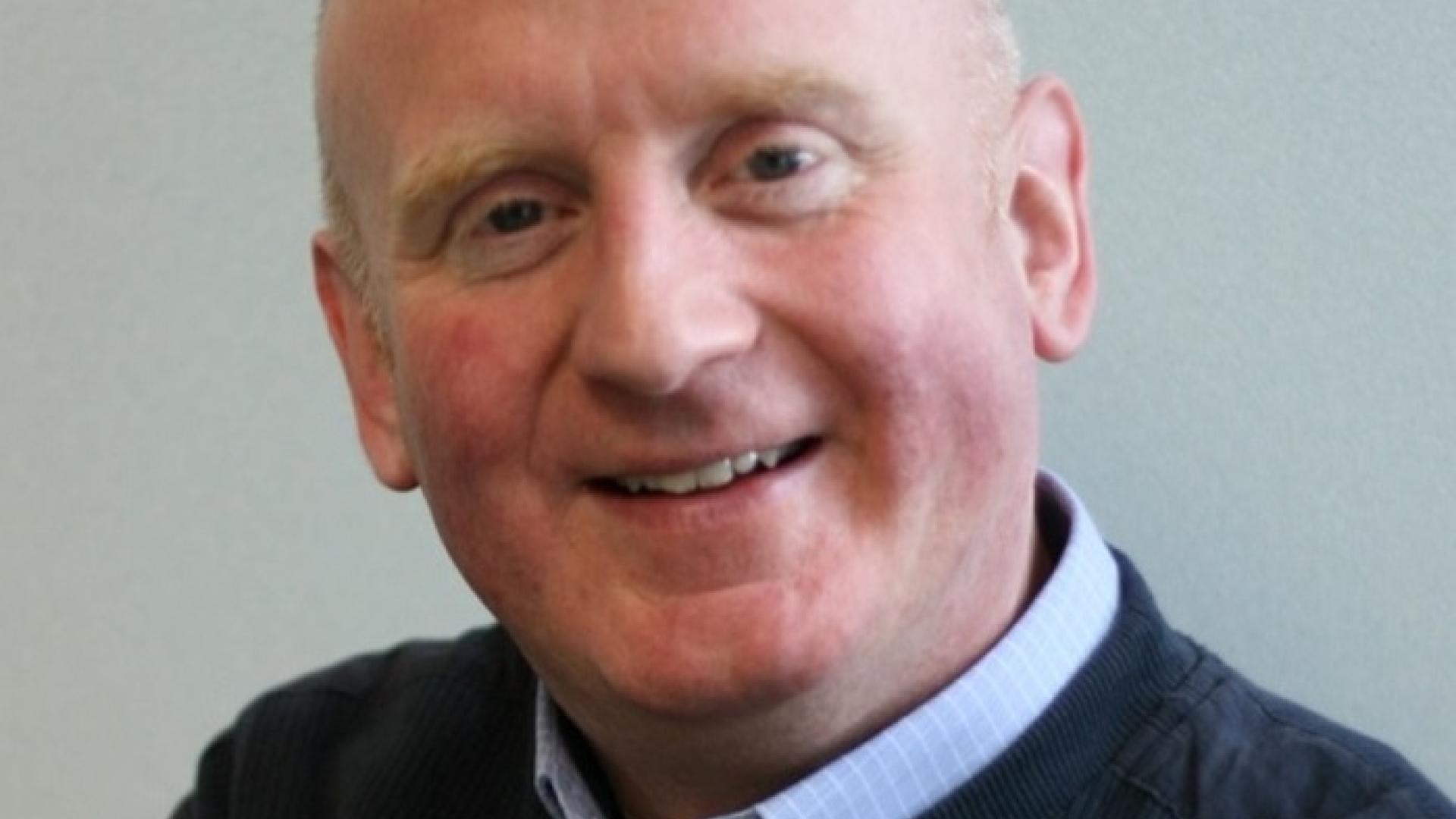 Peter is a white man with a bald head and wearing a pale blue shirt under a navy jumper. He is smiling.