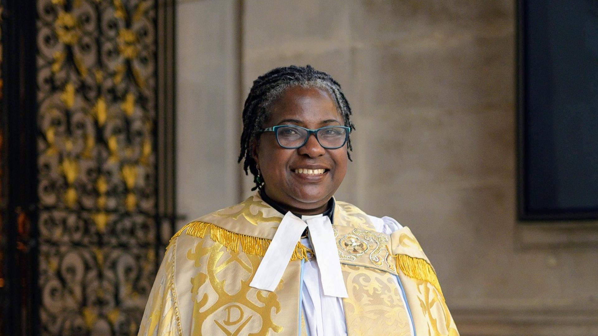 Revd Canon Adéolá is a black woman with short dark hair and glasses. She wears a golden yellow embroidered cope and stands before a portland stone wall and an intricately designed gold and black metal gate.