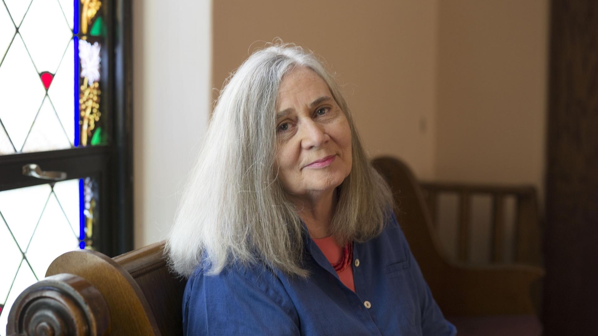 Marilynne is a white woman with long, straight grey hair. She wears a coral pink t-shirt and necklace under a dark blue shirt and sits on a dark wooden pew before a window with stained glass panels. 