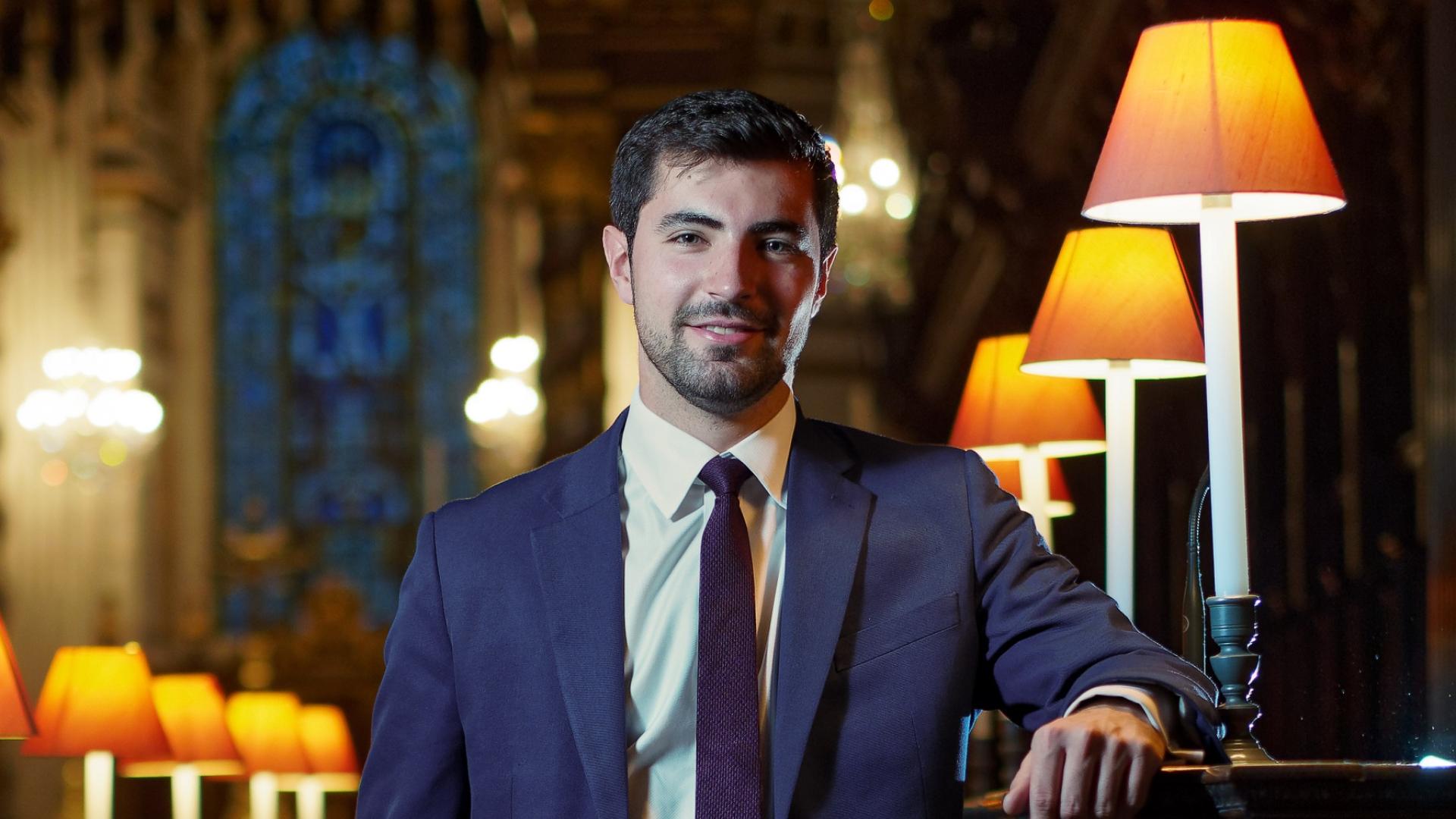 James is a white man with short dark hair wearing a blue suit. He stands in the quire at St Paul's Cathedral, leaning against a stall, with lit lamps alongside.