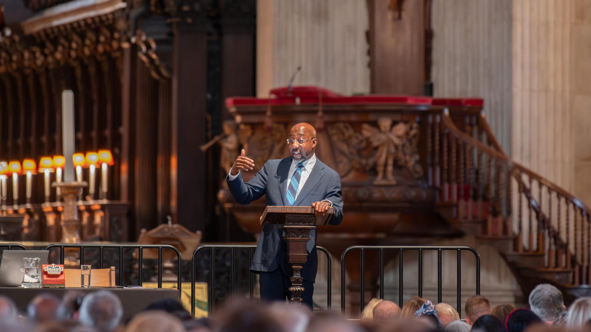 Raphael Warnock, a black man with bald head and glasses wearing a dark suit and blue striped tie, stands a t a lectern on stage in St Paul's Cathedral, the pulpit and quire in the background