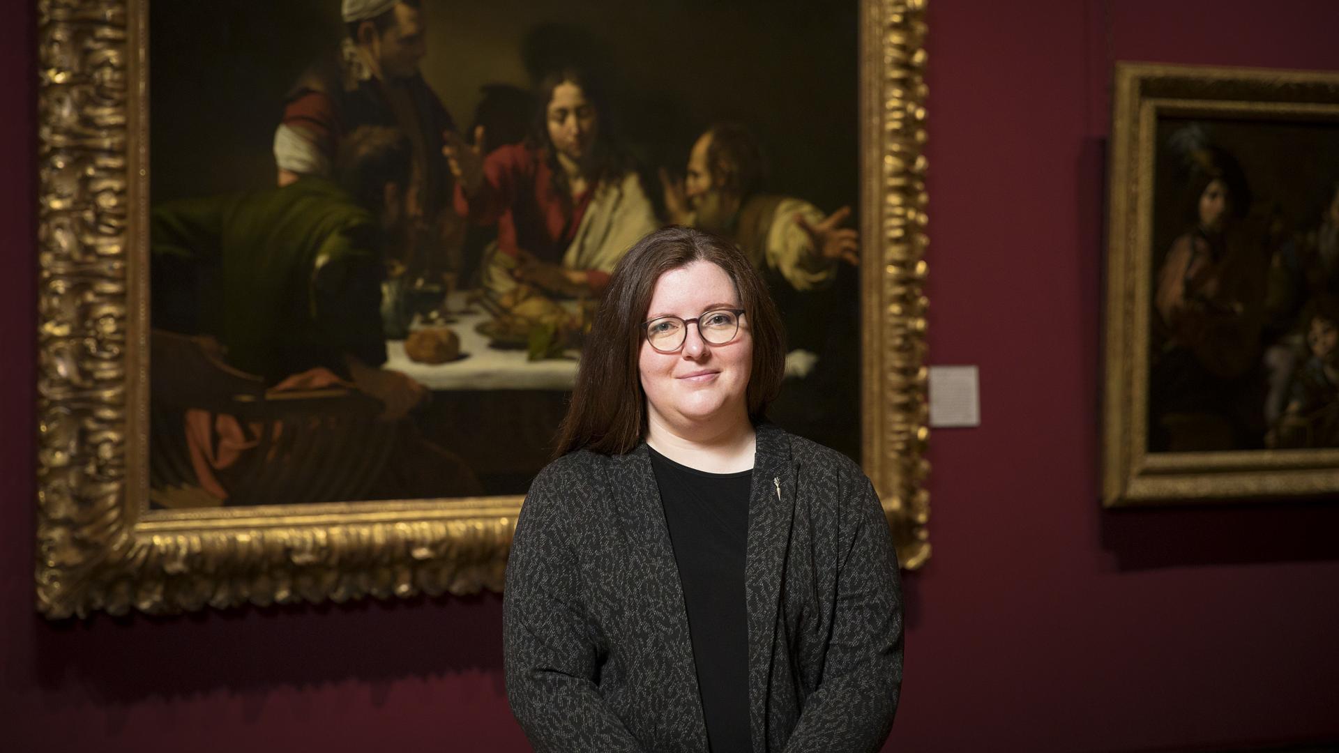 Siobhan Jolley is a white woman with long dark hair and round glasses, wearing a dark grey jacket over a black dress. She stands in the National Gallery in front of Caravaggio’s painting The Supper at Emmaus.