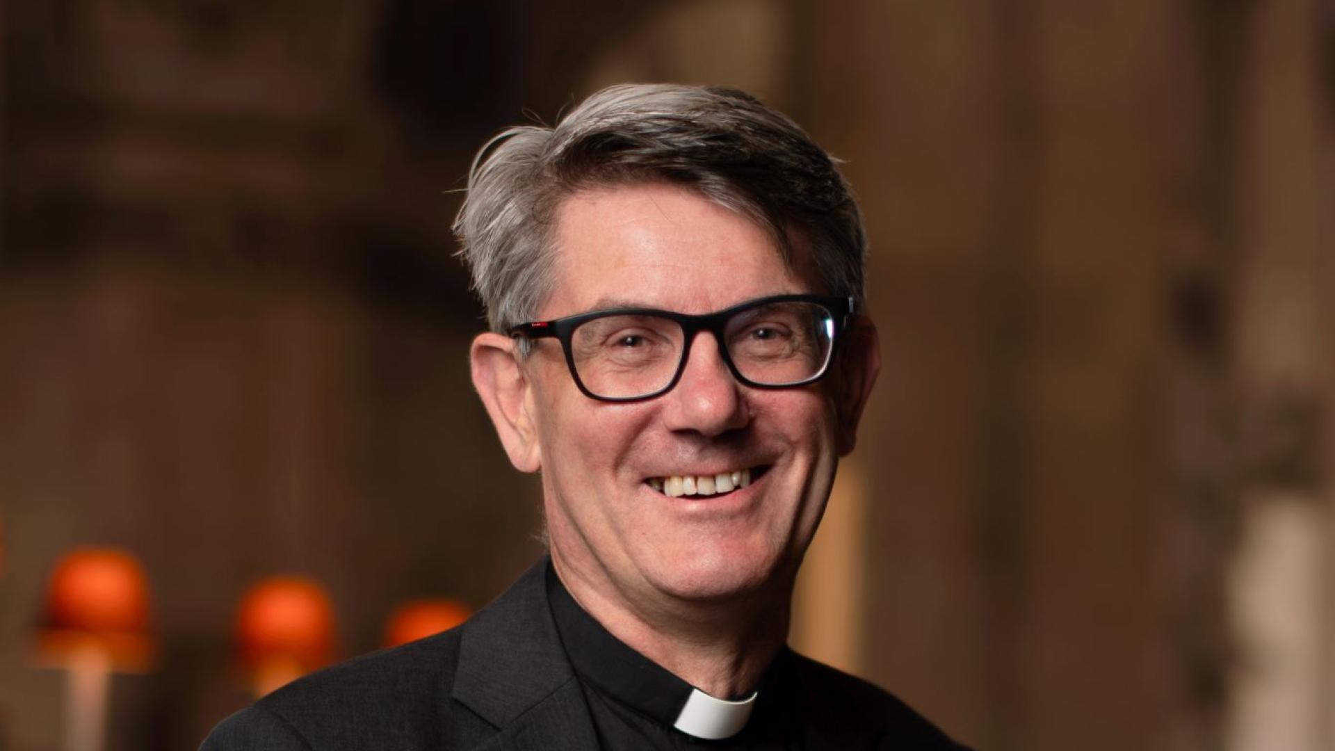 Andrew is a white man with short grey hair and black rimmed glasses wearing a clerical collar, black shirt and black jacket. The lamps of the quire of St Paul's in the background.