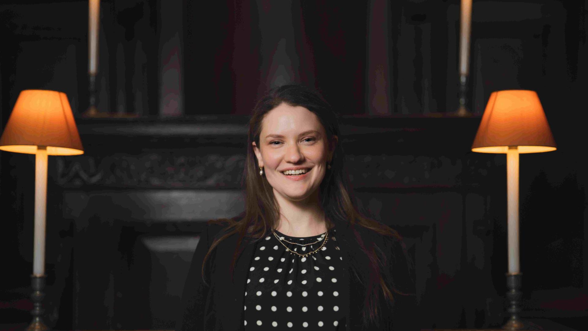 A photo of Hilary Punnet, Assistant Director of Music at St Paul's Cathedral. Hilary is a white woman with long brown hair. She is smiling at the camera, and wearing a black polka dot dress with a black blazer.
