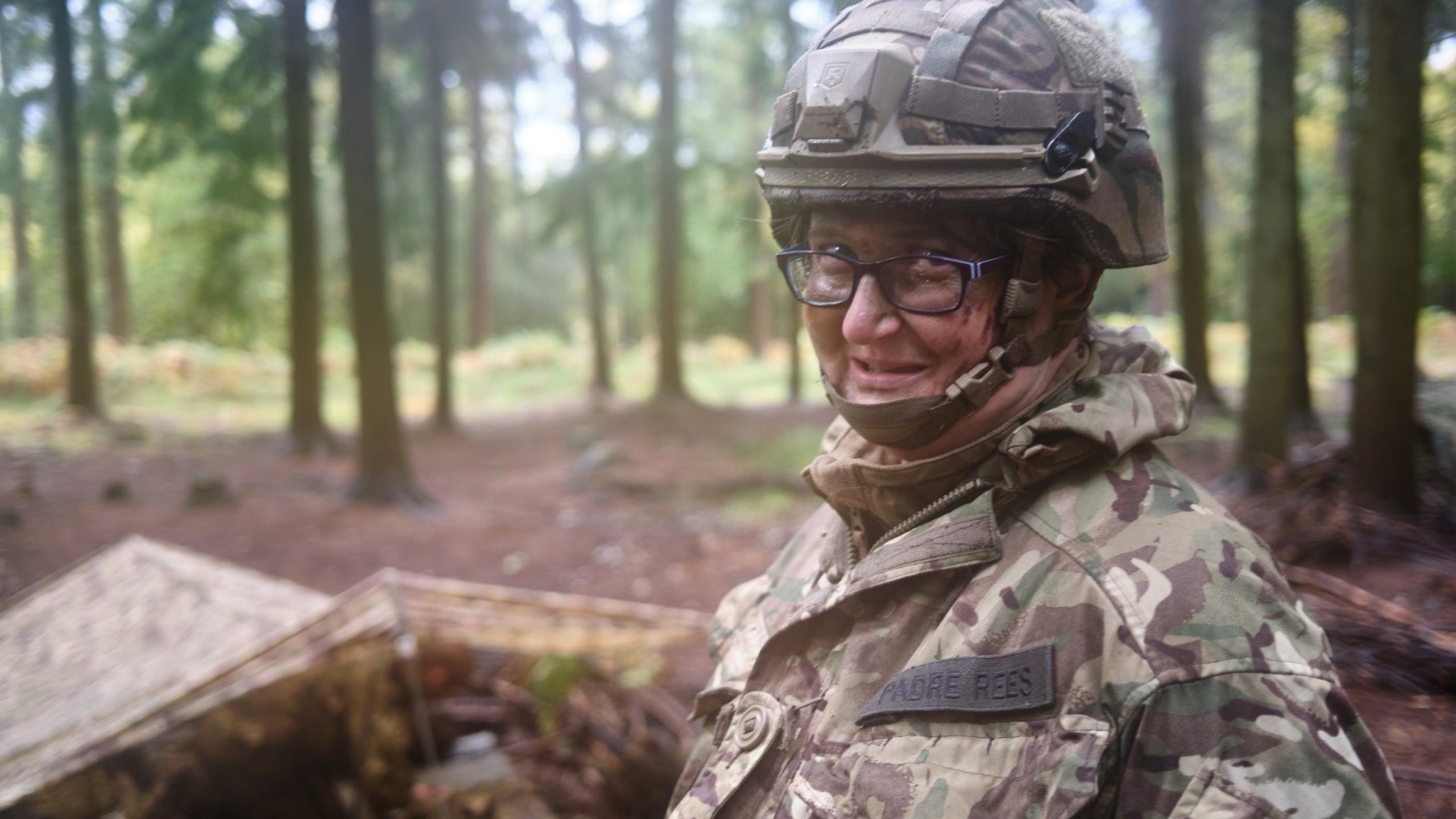 Adele wears army fatigues and a helmet and stands in front of a shelter in woodland. She wears a badge which says Padre Rees.