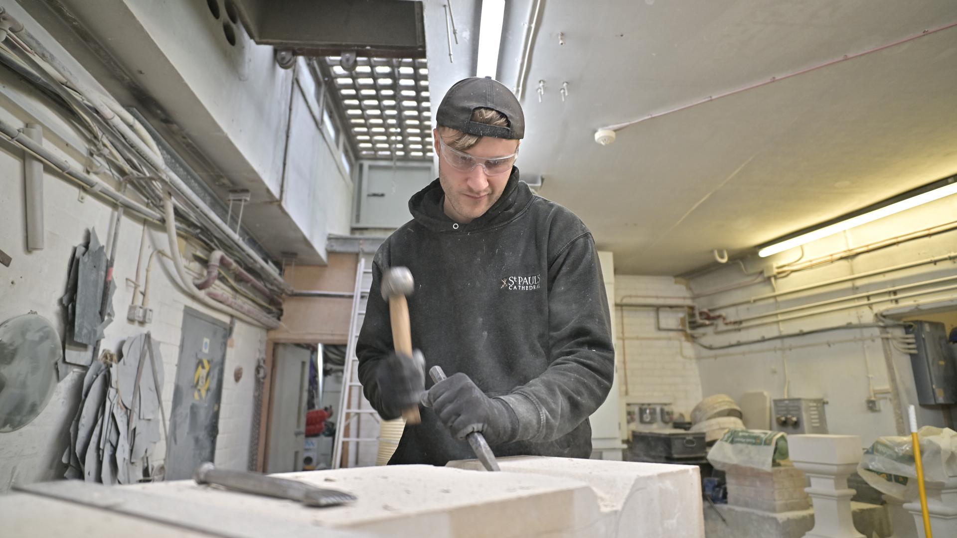 male person wearing a cap and black clothes in a workshop using a hammer and chisel to carve a stone