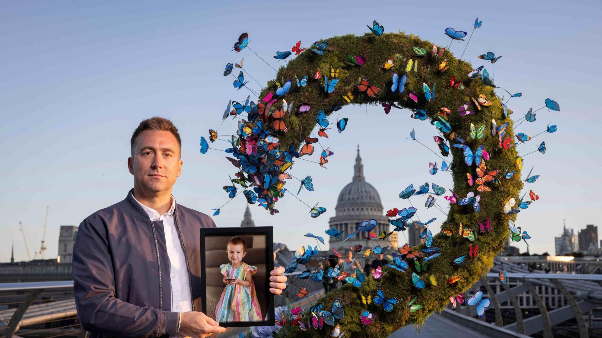 A man stands on Millennium Bridge in London, dressed in a white shirt and blue jacket, with short blond hair. He is holding a photo of his late daughter, who passed away from cancer. He is standing beside an art work for Great Ormond Street Hospital Charity, made of butterflies, framing the dome of St Paul's Cathedral.