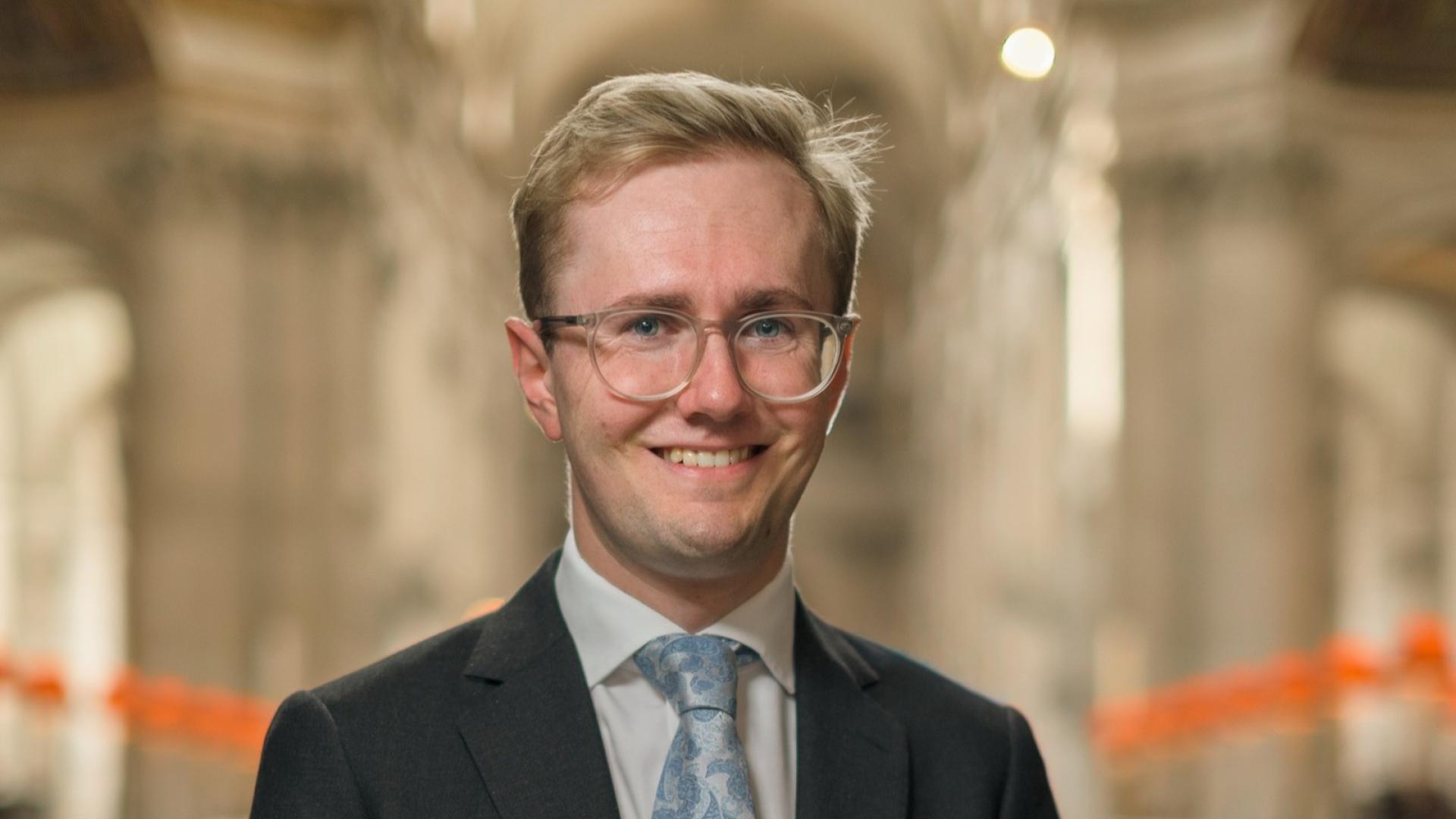 Hamish Wagstaff smiling, wearing glasses, dark suit, and light blue tie with floral pattern