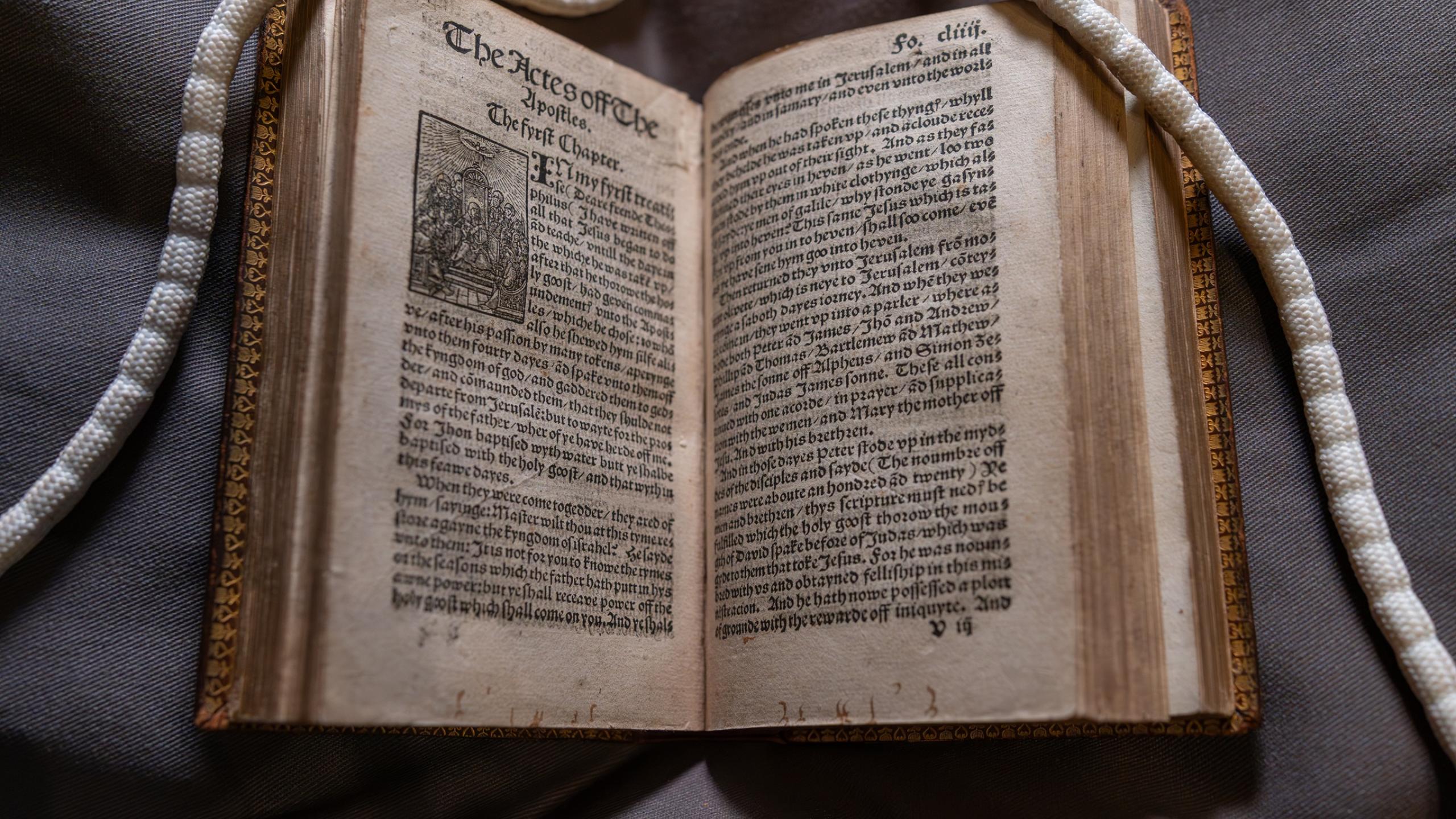 Tyndale Bible open at "The Acts of the Apostles". Long white "snake" weights are visible holding the top right and left corners of paper open. 