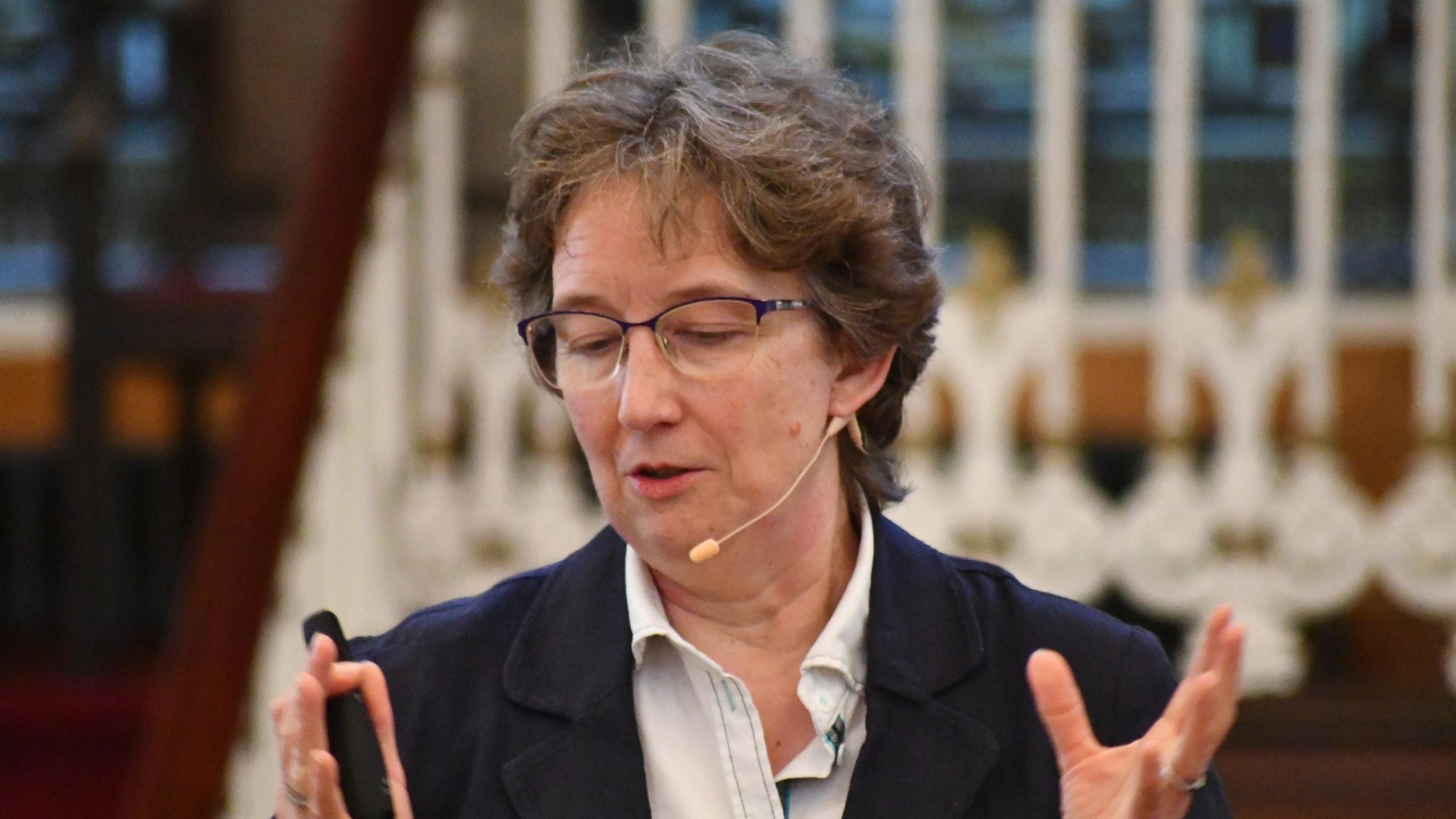 Helen has short brown hair and glasses, and stands with hands raised and wearing an ear microphone as she talks.