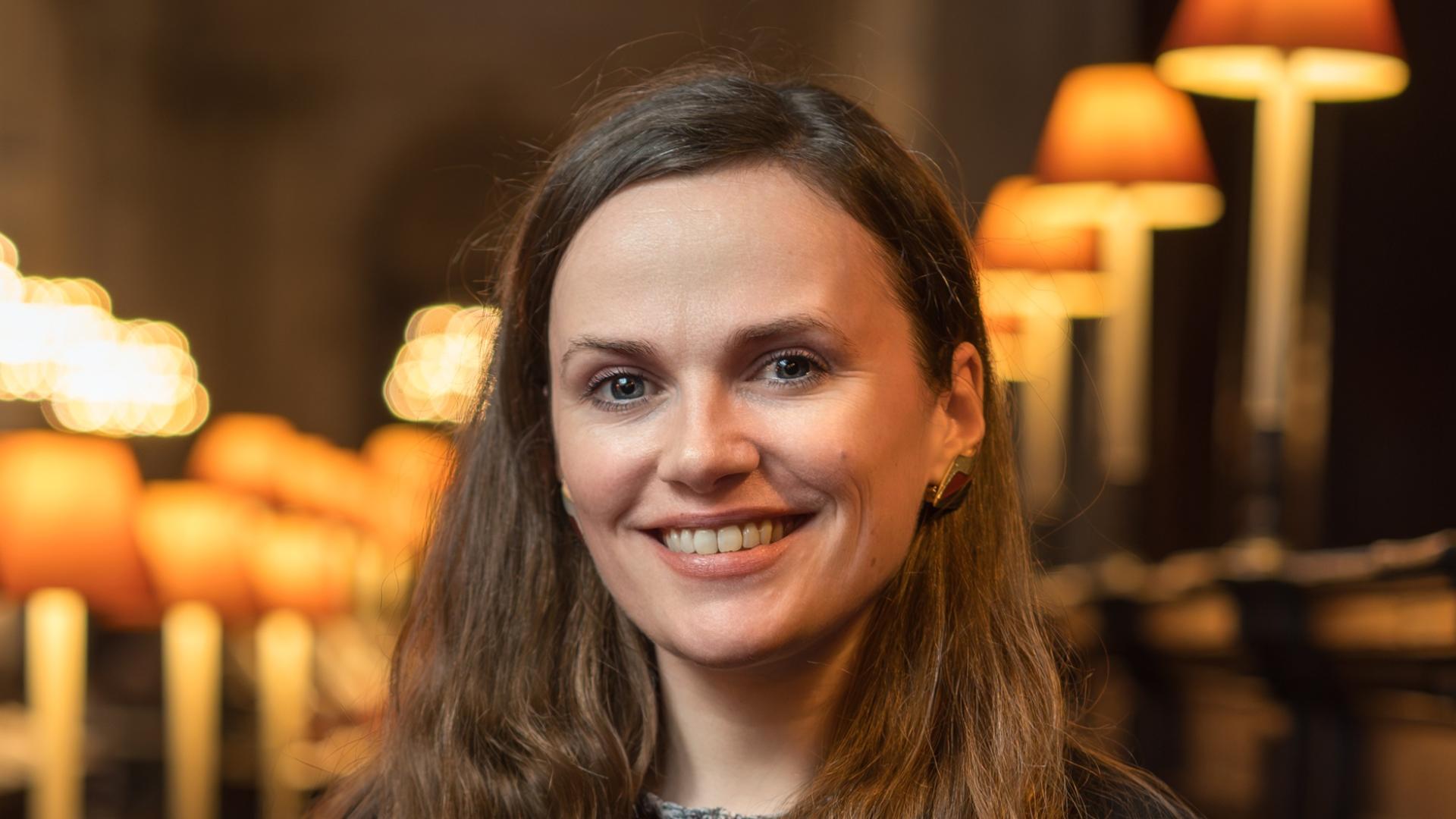 Laurel has long brown hair and stands in the quire of St Paul's Cathedral with lit lamps behind her