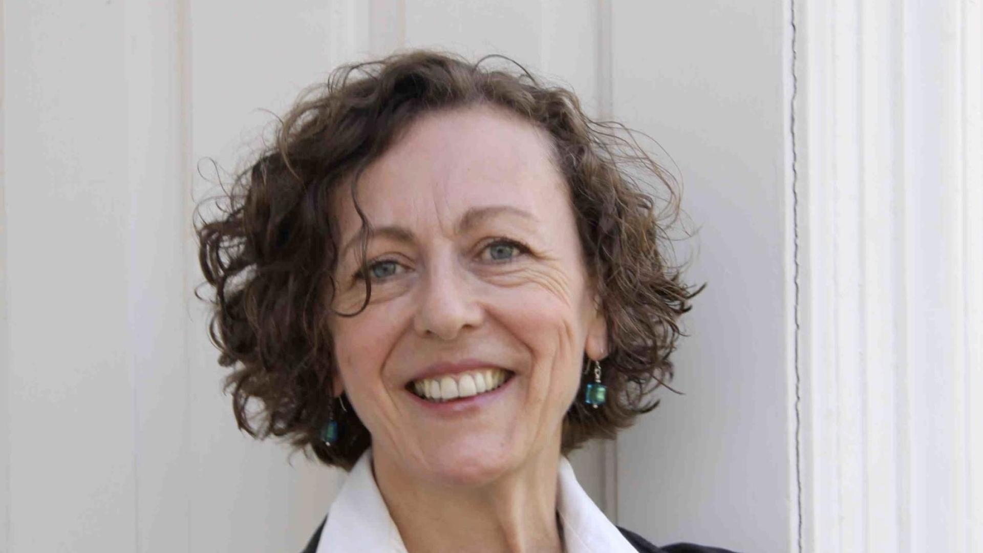 Maggi has short wavy brown hair and wears a white shirt under a black jacket