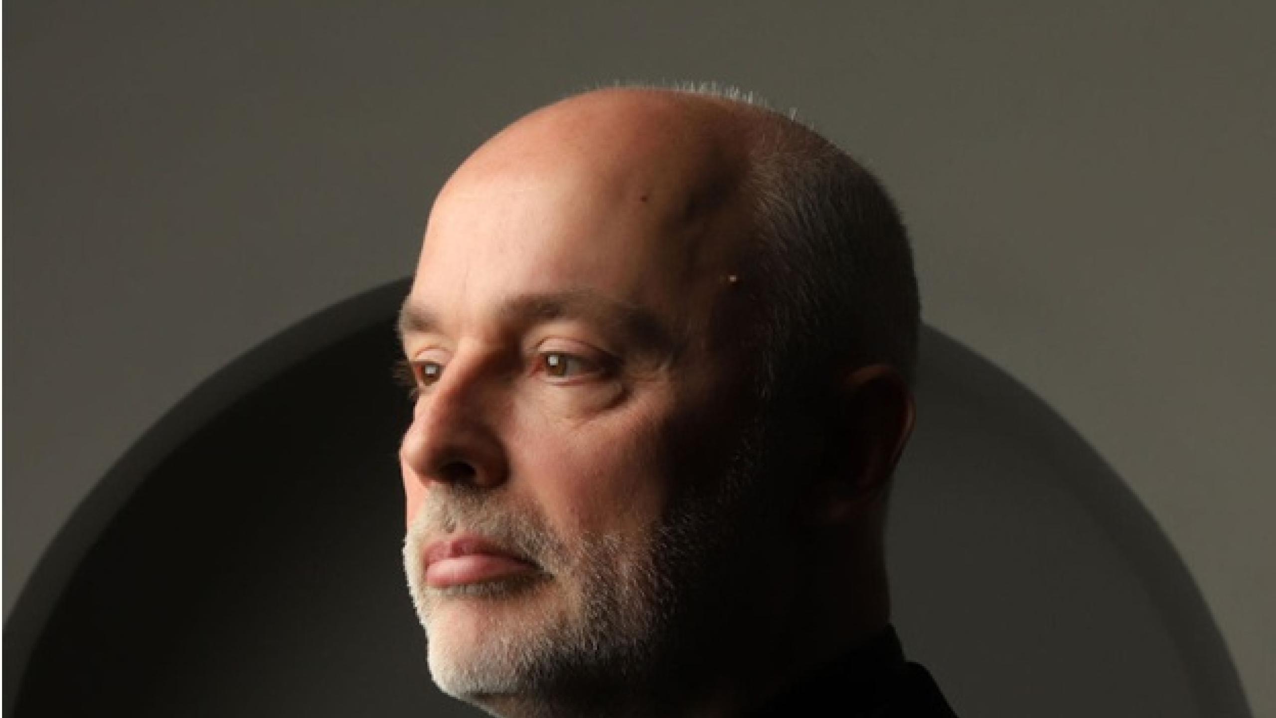 Patrick has short grey hair and a grey beard, wears a black suit and shirt and faces to the left