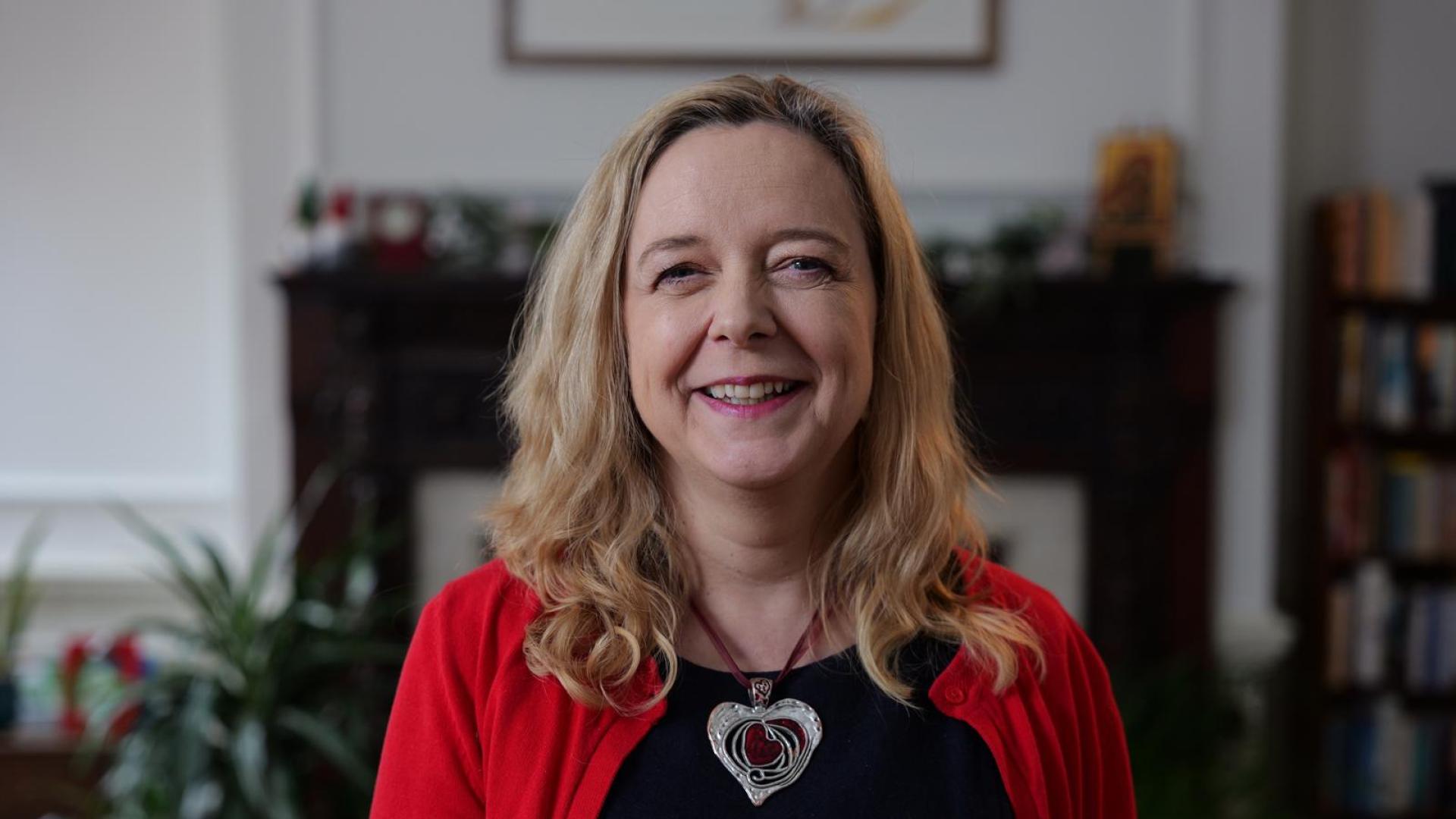 Paula wears a red cardigan over a black dress and stands in front of  large fireplace