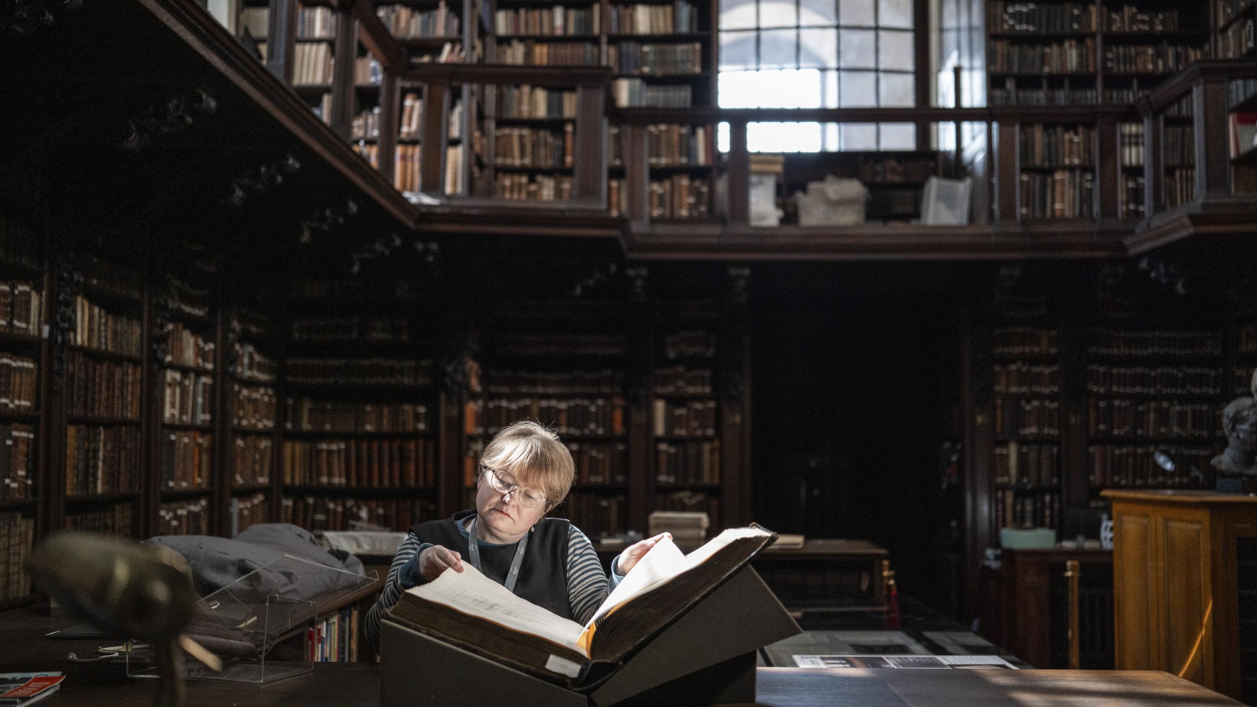 The Library of St Paul's Cathedral. Librarian Anna is reading a manuscript, lit by a sunbeam from the arched window behind.