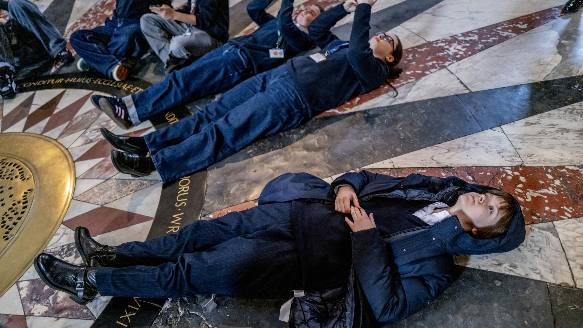 School children lying on a marble floor pointing up towards the ceiling