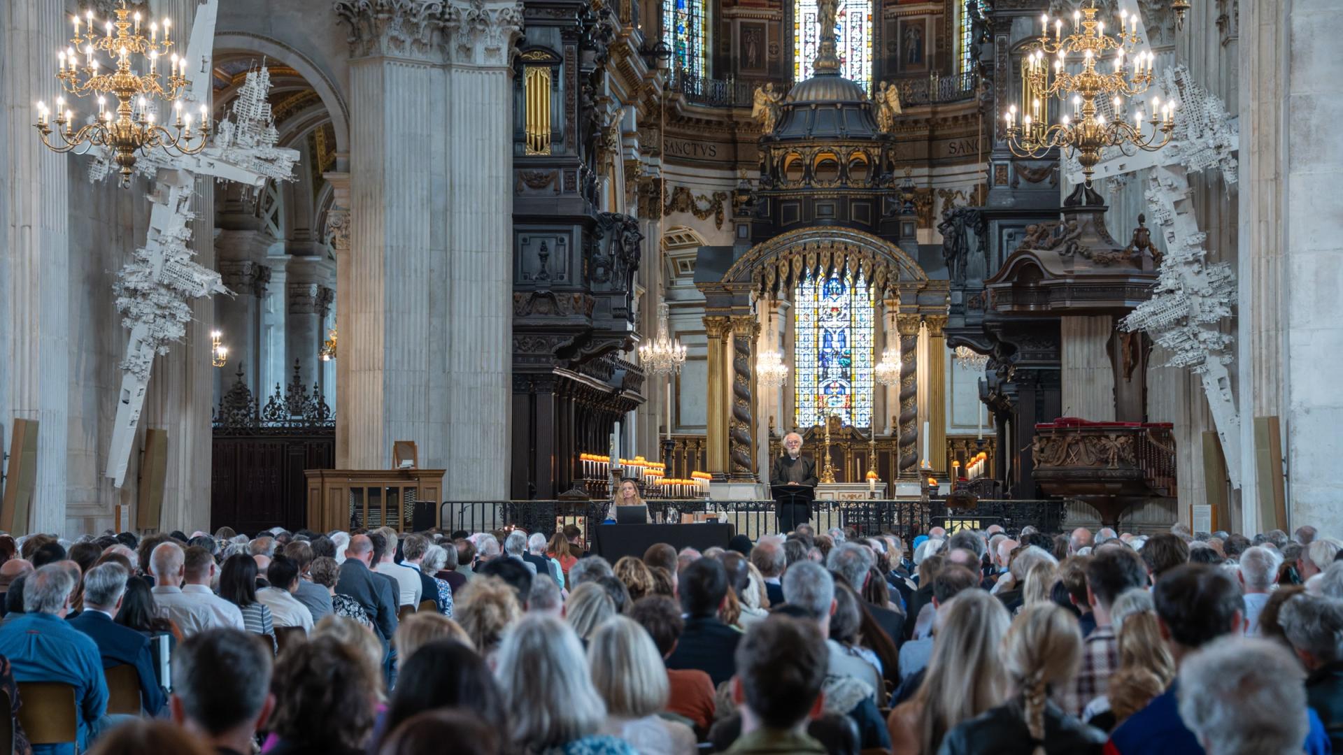 A large audience in the nave of the cathedral with the quire and high altar in the background and a stage visible with two speakers