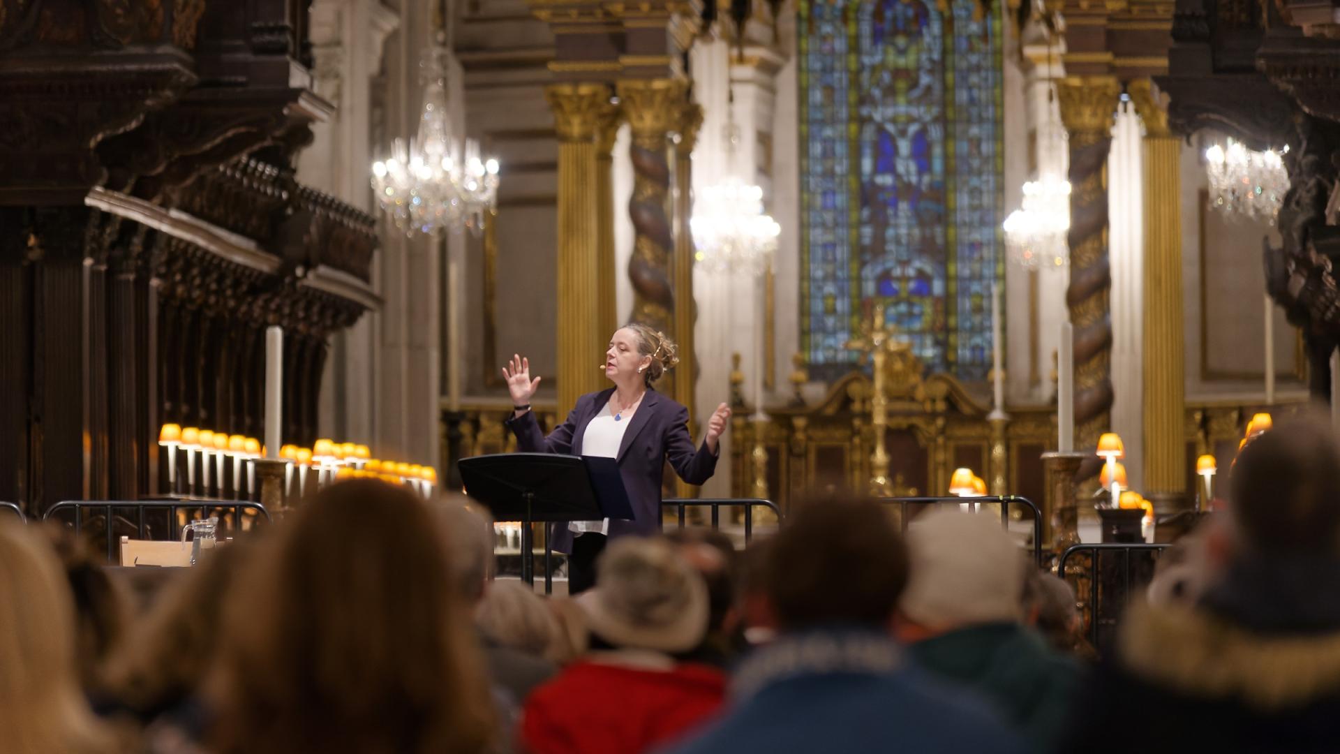 Paula Gooder stands on a stage with hands raised as she talks with a large audience in front and the quire and high altar in the back