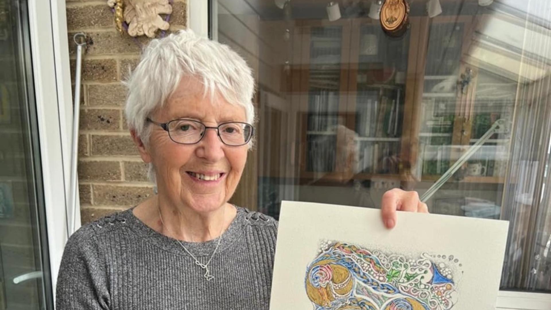 A woman with short white hair and glasses standing in a conservatory holding a finished piece of art decorated with gold leaf and pastel colours