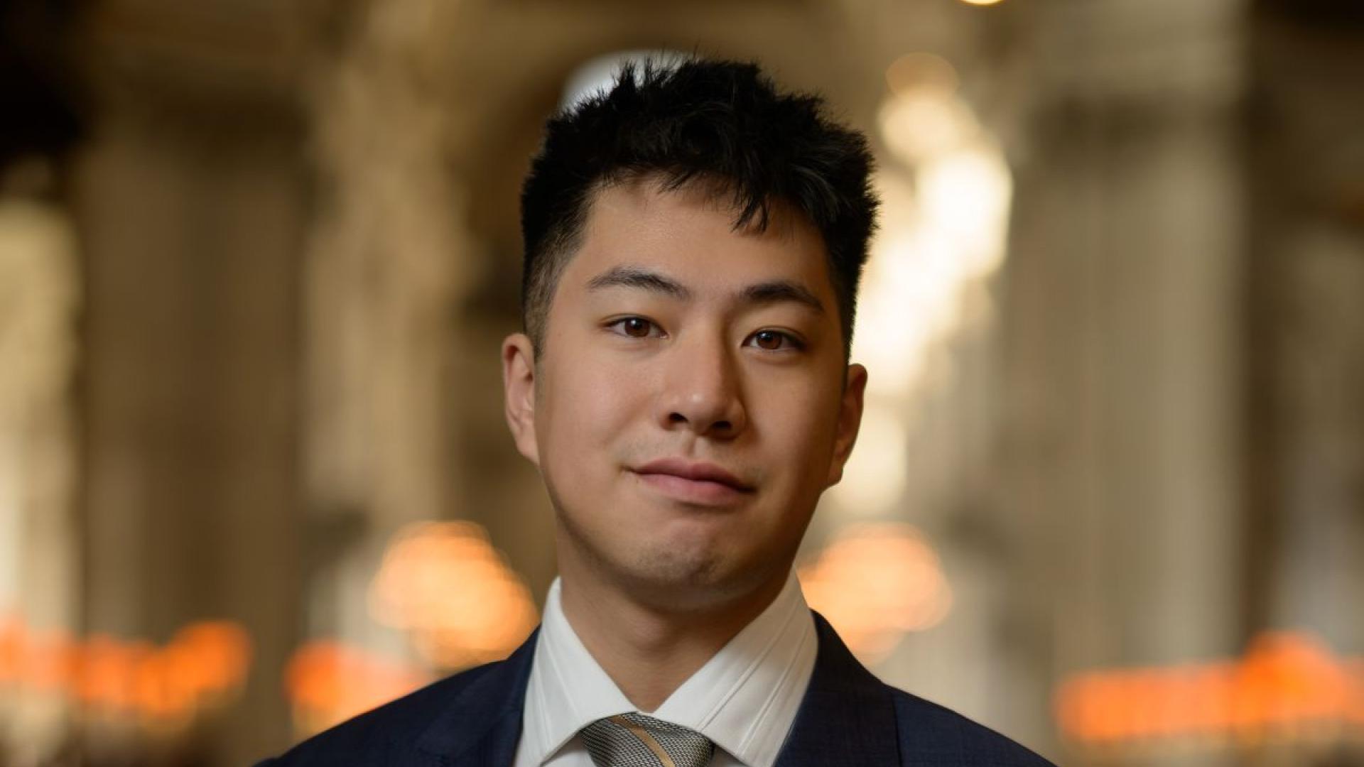 Jeremy Ng smiling wearing a navy blue suit jacket, white shirt, and grey tie with black and gold diagonal stripes