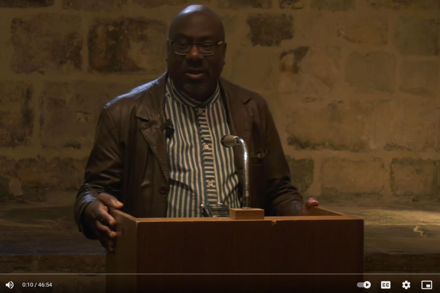 A photo taken from the film of Anthony Reddie speaking in the Wren Suite at St Paul's Cathedral. Pictured standing at a lectern in front of a wall of exposed brickwork, he is a Black man wearing a striped shirt and a brown leather jacket.