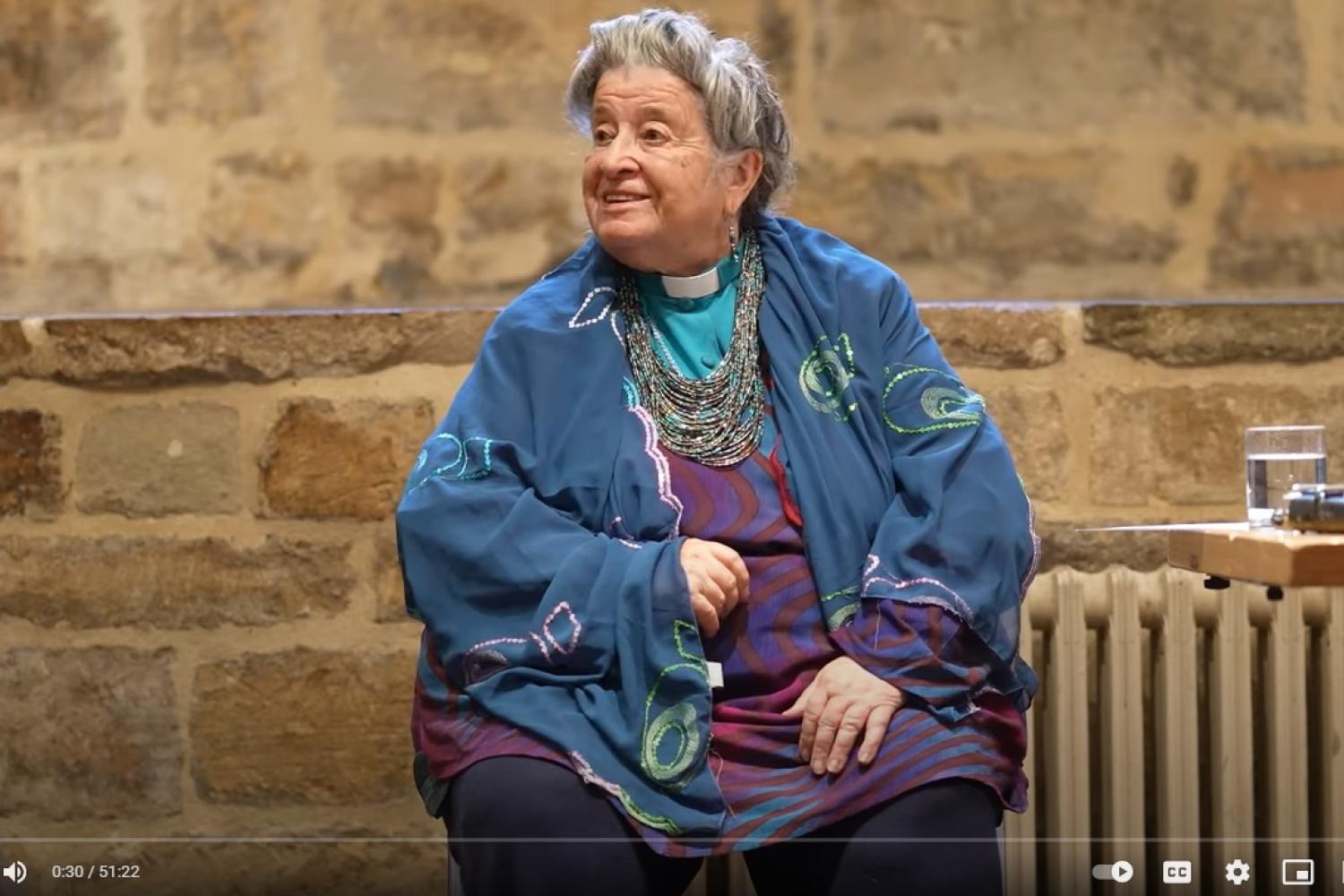 June Boyce-Tillman is a white woman with grey hair gathered in a bun, wearing a turquoise blue clerical shirt under a blue shawl. She is pictured sitting indoors in front of an exposed brick wall.
