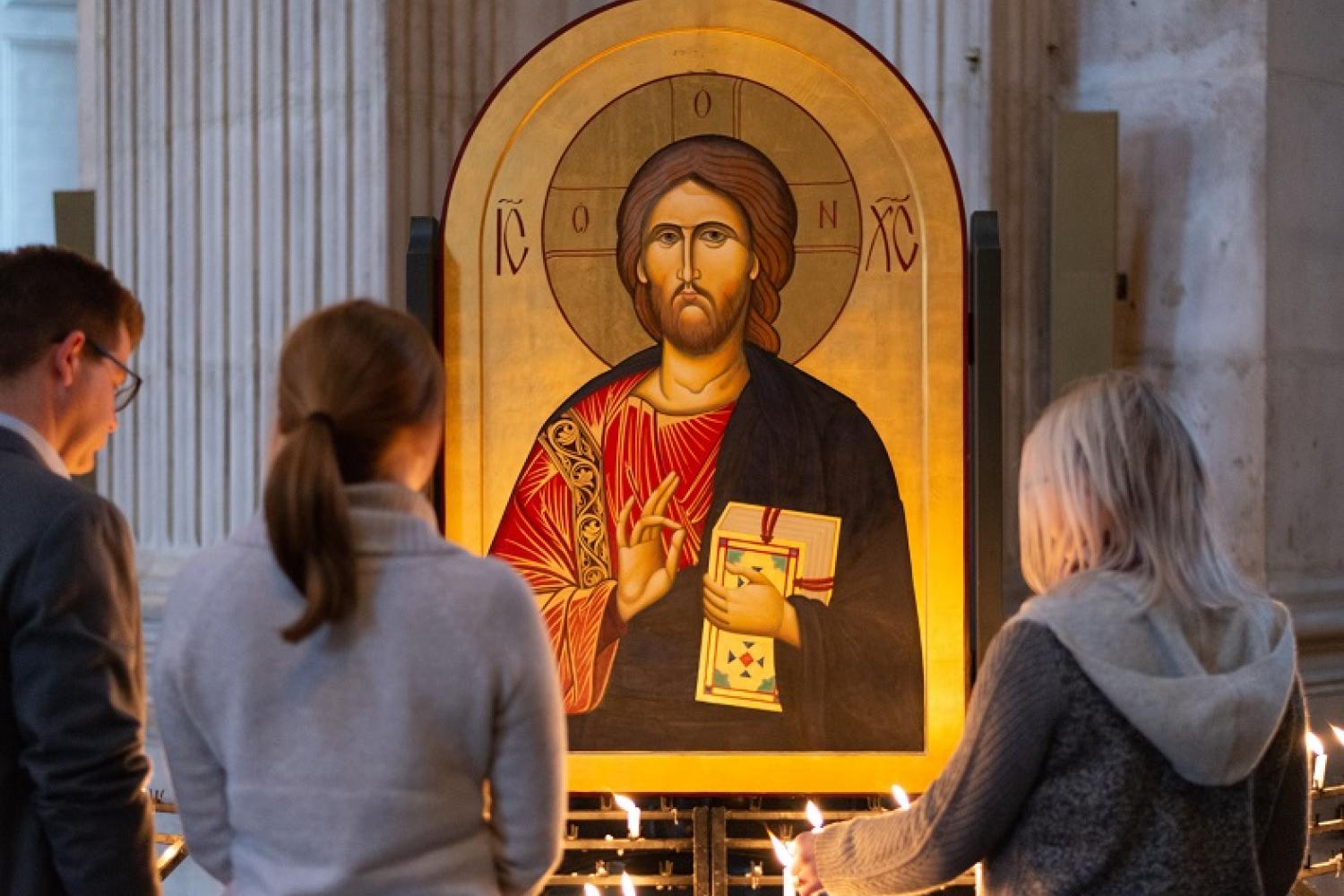 Two women and a man stand before an Icon of Jesus and light candles as they reflect.