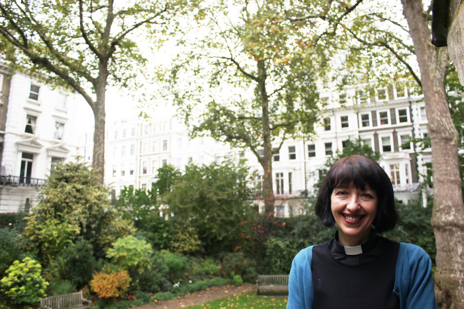 Carys, a white woman with short brown hair wearing a clerical collar, stands in a garden square in a city