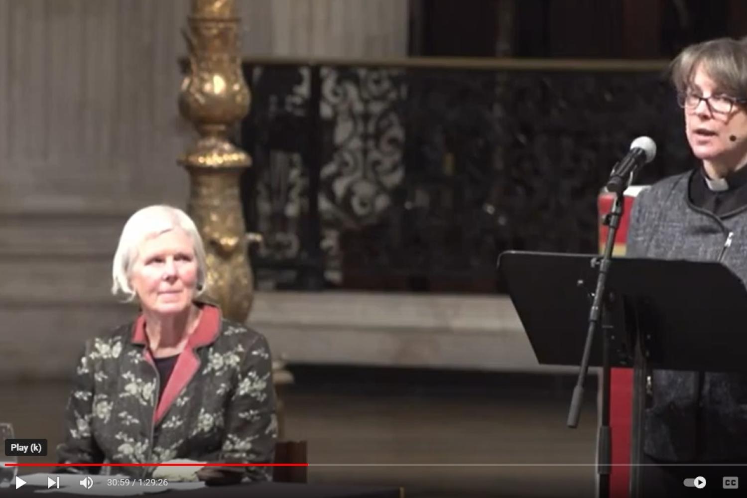 Lucy Winkett speaking from the lectern at St Paul's Cathedral with Marian Partington listening from a table beside her.