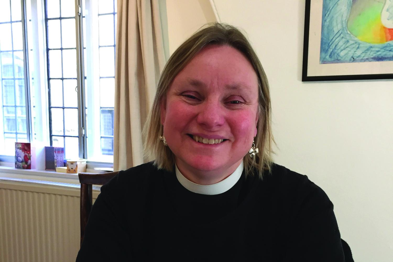 Jessica Martin is a white woman with short blond hair, photographed in clerical shirt and in her house, sitting leaning on a table and smiling.