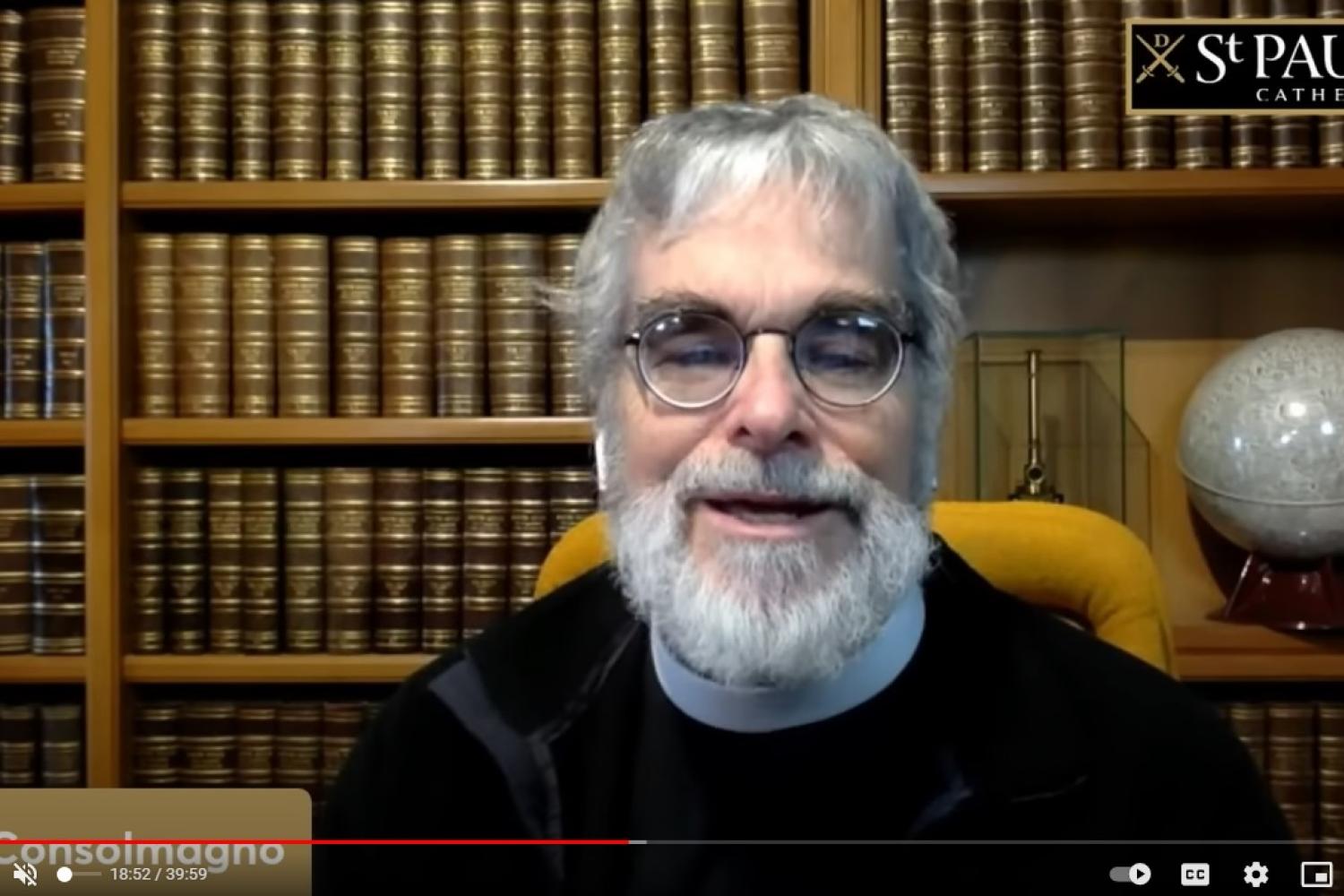Brother Guy is a white man with round glasses, grey hair and a full grey beard sitting in front of bookshelves full of bound leather books., and a globe behind to his left.