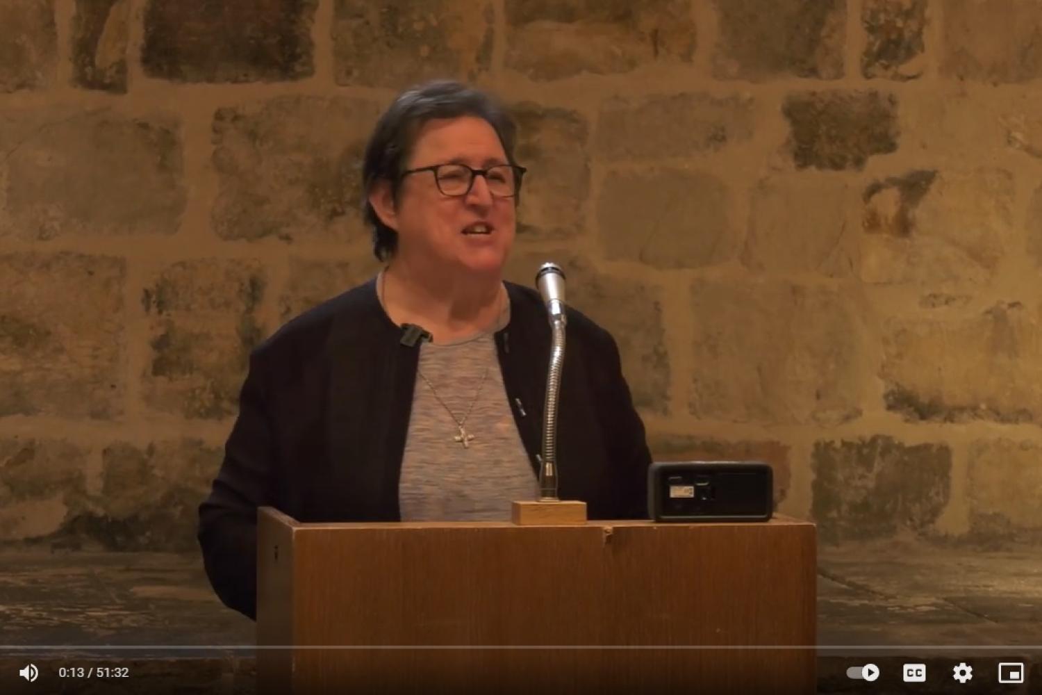 Gemma Simmonds, a white woman with glasses and short brown, speaking from the lectern in the Wren Suite, an exposed brick wall behind her.