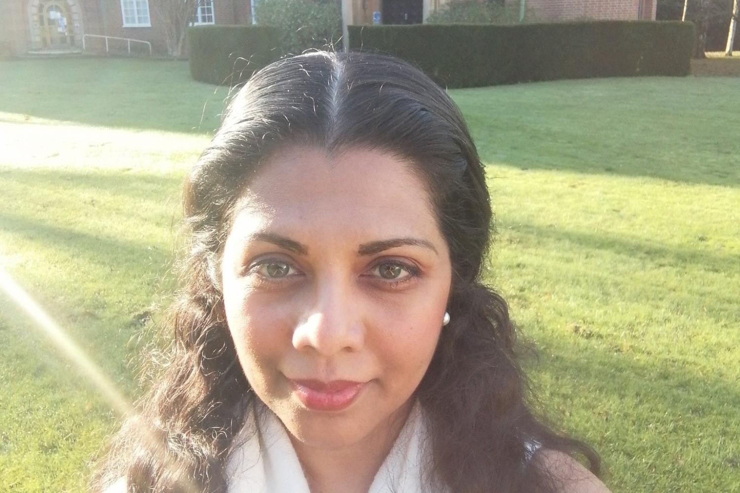 Sanjee Perera is a young Asian woman with long dark hair, photographed on a green lawn with sunlight across the lawn behind her, and a chapel in the background.