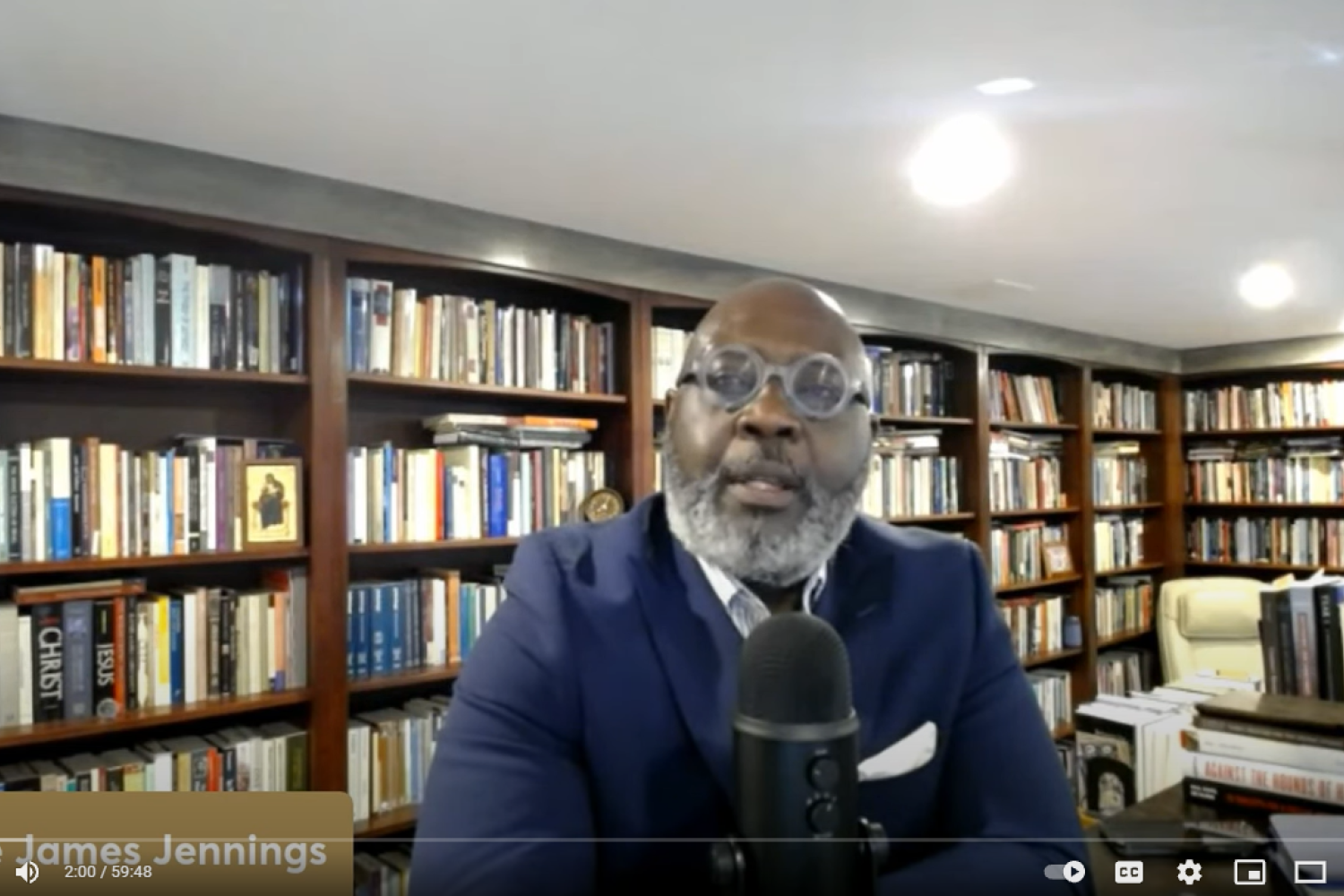 Willie Jennings on screen during the lecture in his office with rows of bookcases behind