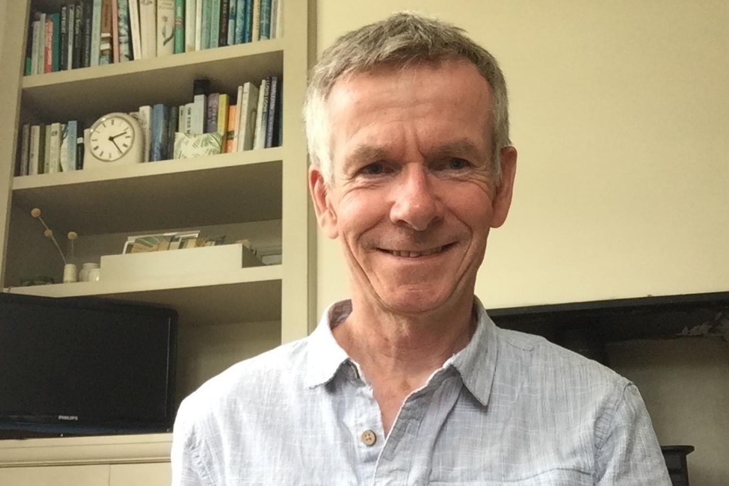 Christopher Chapman is a white man with short grey hair wearing a pair grey casual shirt and sitting in front of a bookcase, cupboard and fireplace.