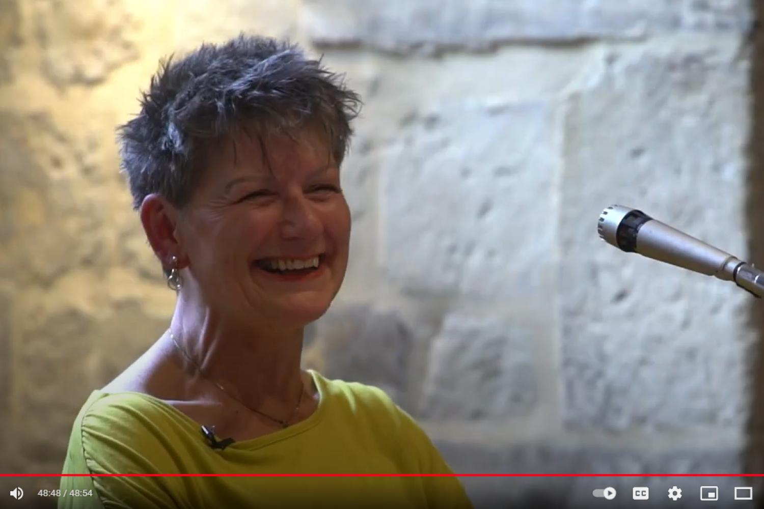 Claire Gilbert is a white woman with short grey hair wearing a yellow sweater and smiling to the audience. She stands in front of the sunlit alcove in the Wren Suite of the Cathedral.