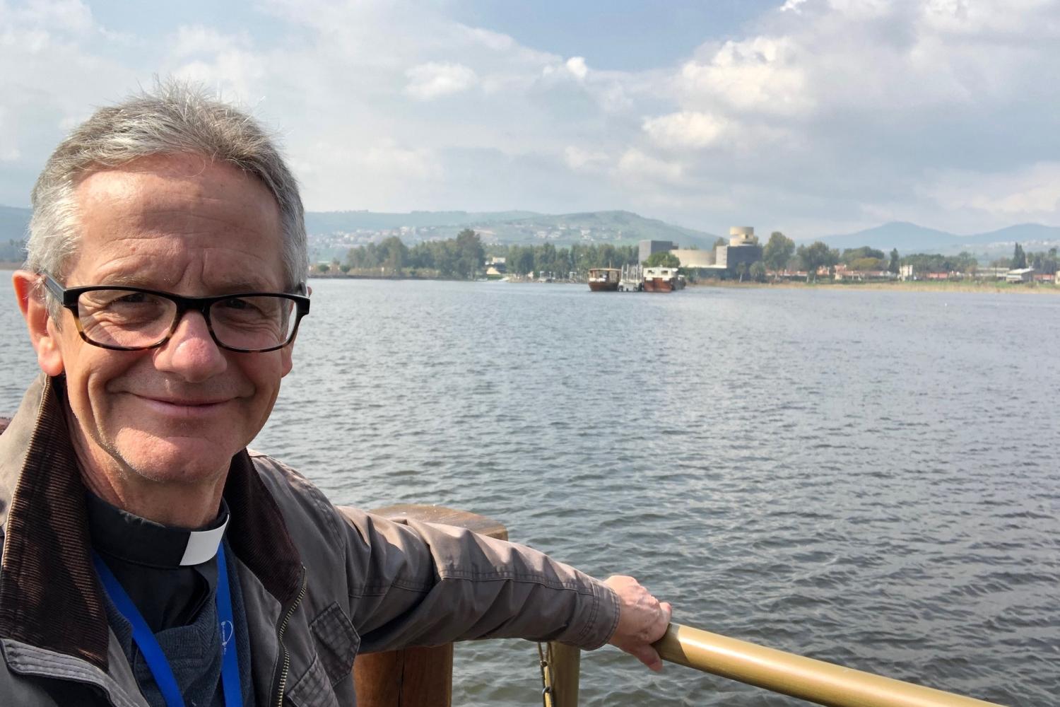 Richard is a white man with short grey hair and black rimmed glasses. He wears a clerical collar and black shirt under a casual light jacket and stands holding onto the wooden railing of a boat or a jetty on a lake. The shoreline in the distance has trees, and some buildings, and there are mountains in the distant background. The sky is pale blue with some clouds and the sun is shining on Richard.