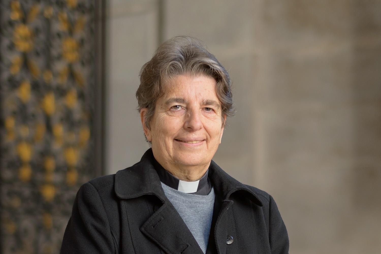 Paula is a white woman with short dark grey hair and wears a clerical collar under a grey shirt and black double breasted coat. 
