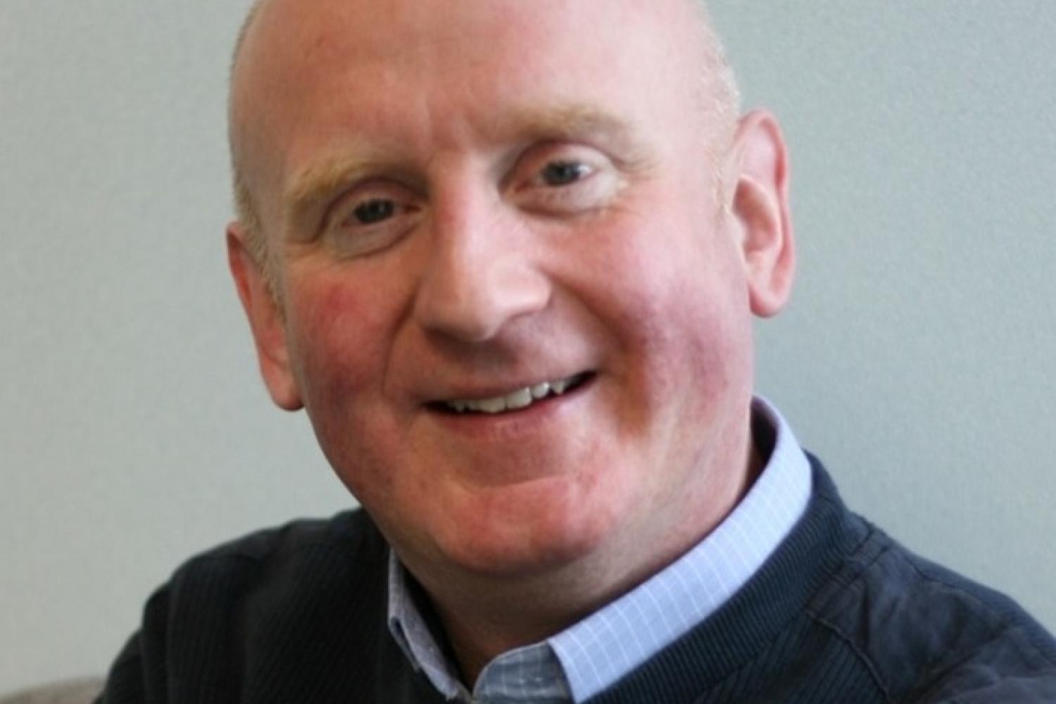 Peter is a white man with a bald head and wearing a pale blue shirt under a navy jumper. He is smiling.