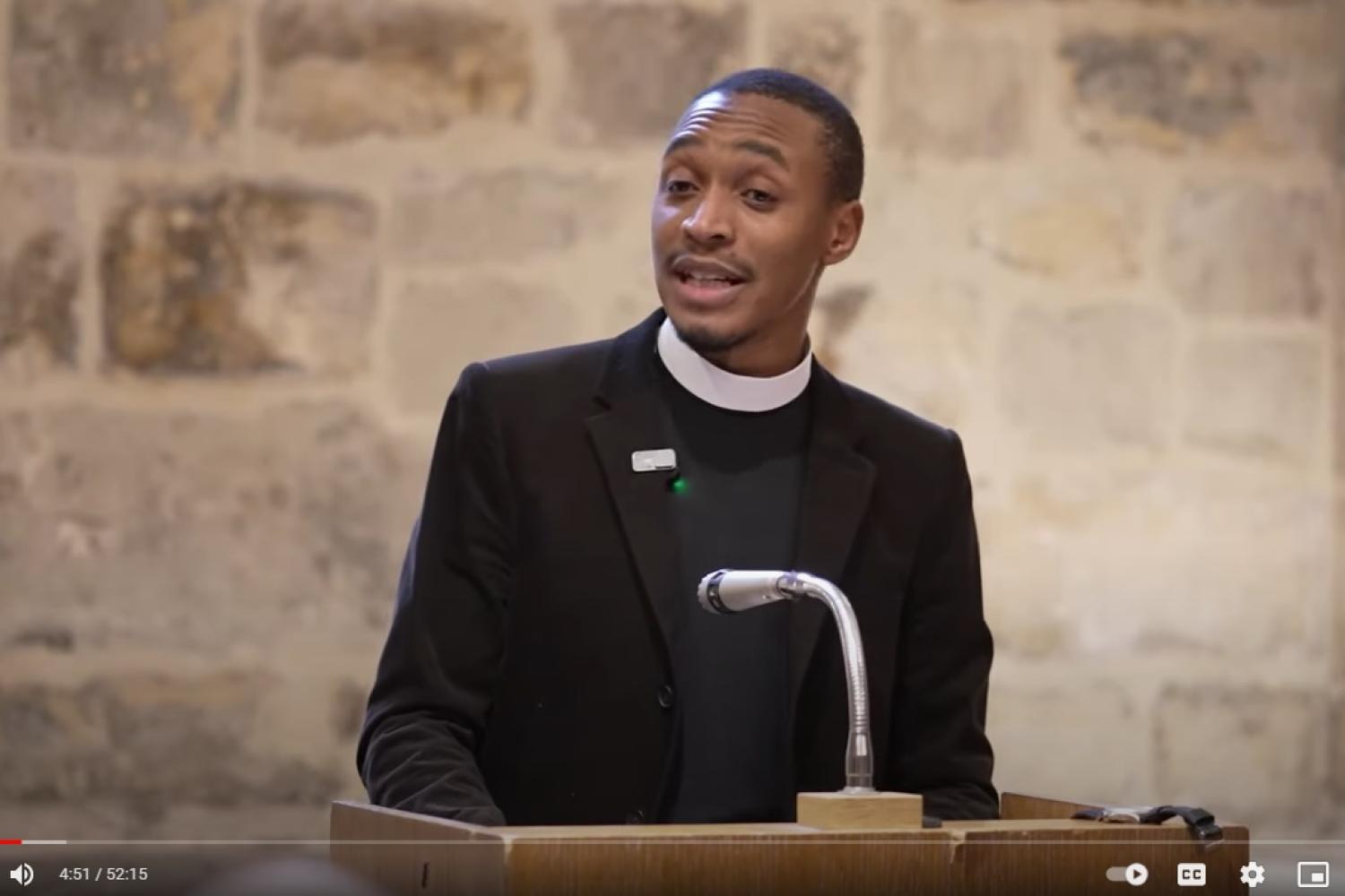 Jarel is a young black man wearing a clerical collar under a black shirt and suit, and he is standing at a lectern talking, in a room with an old exposed stone wall behind him.