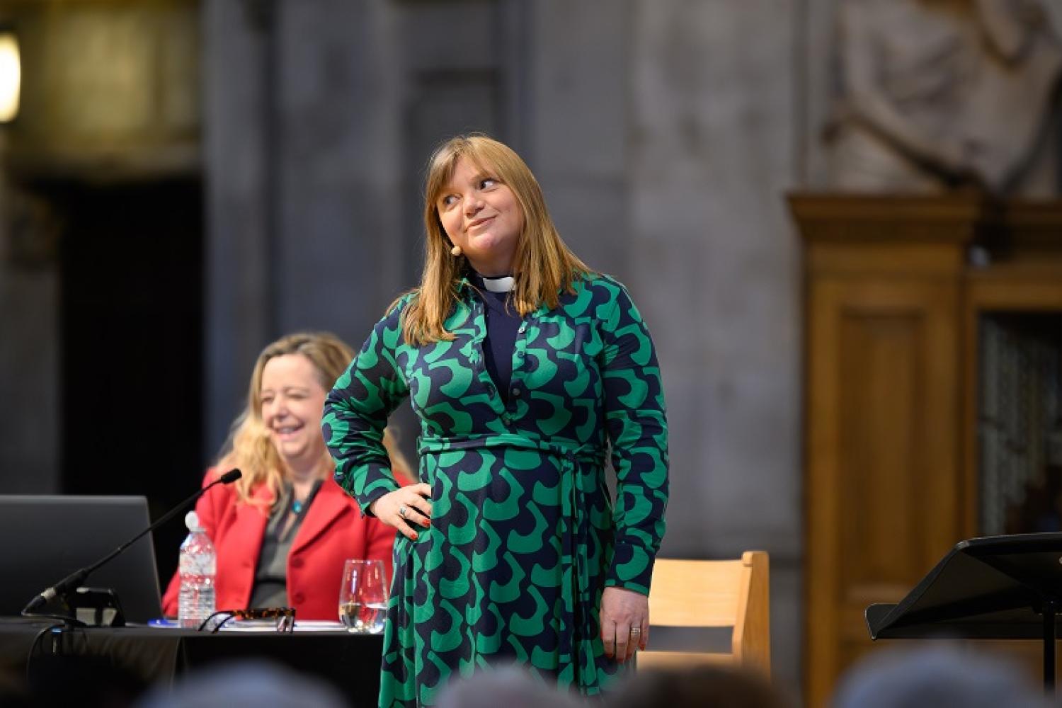 Kate on stage wearing a green dress and clerical collar, with her hand on her hip, giving side eye to the audience as Paula laughs in the background.
