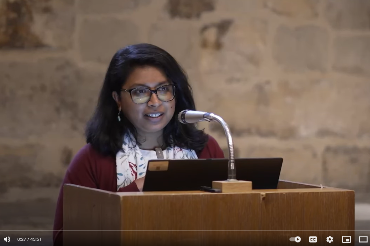 Anupama is a your Asian woman with shoulder length black hair wearing glasses. She is standing at a lectern with a microphone in the Wren Suite at St Paul's Cathedral.